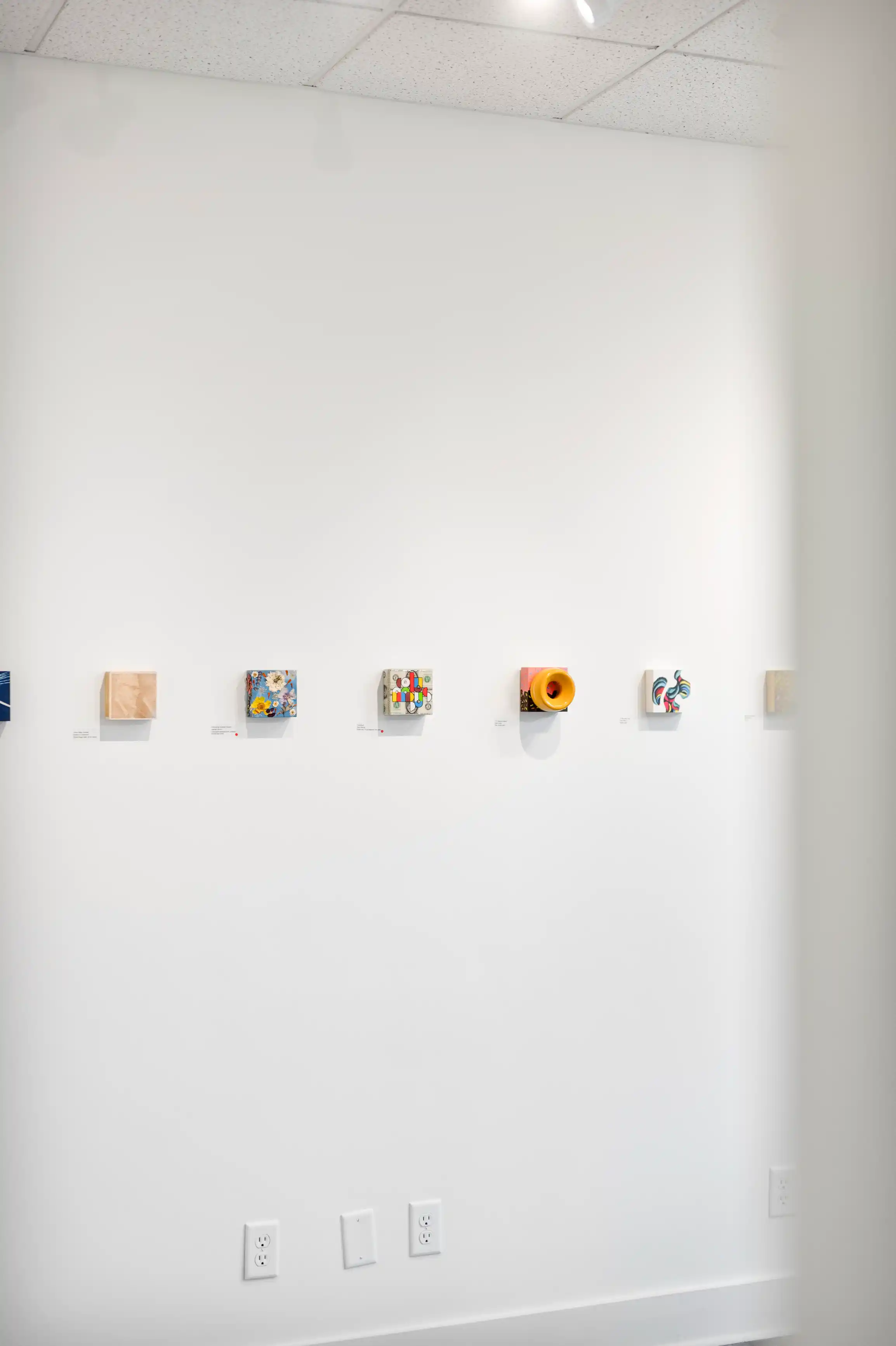 Art installation on a white gallery wall featuring six colorful abstract pieces, each accompanied by a descriptive label below.