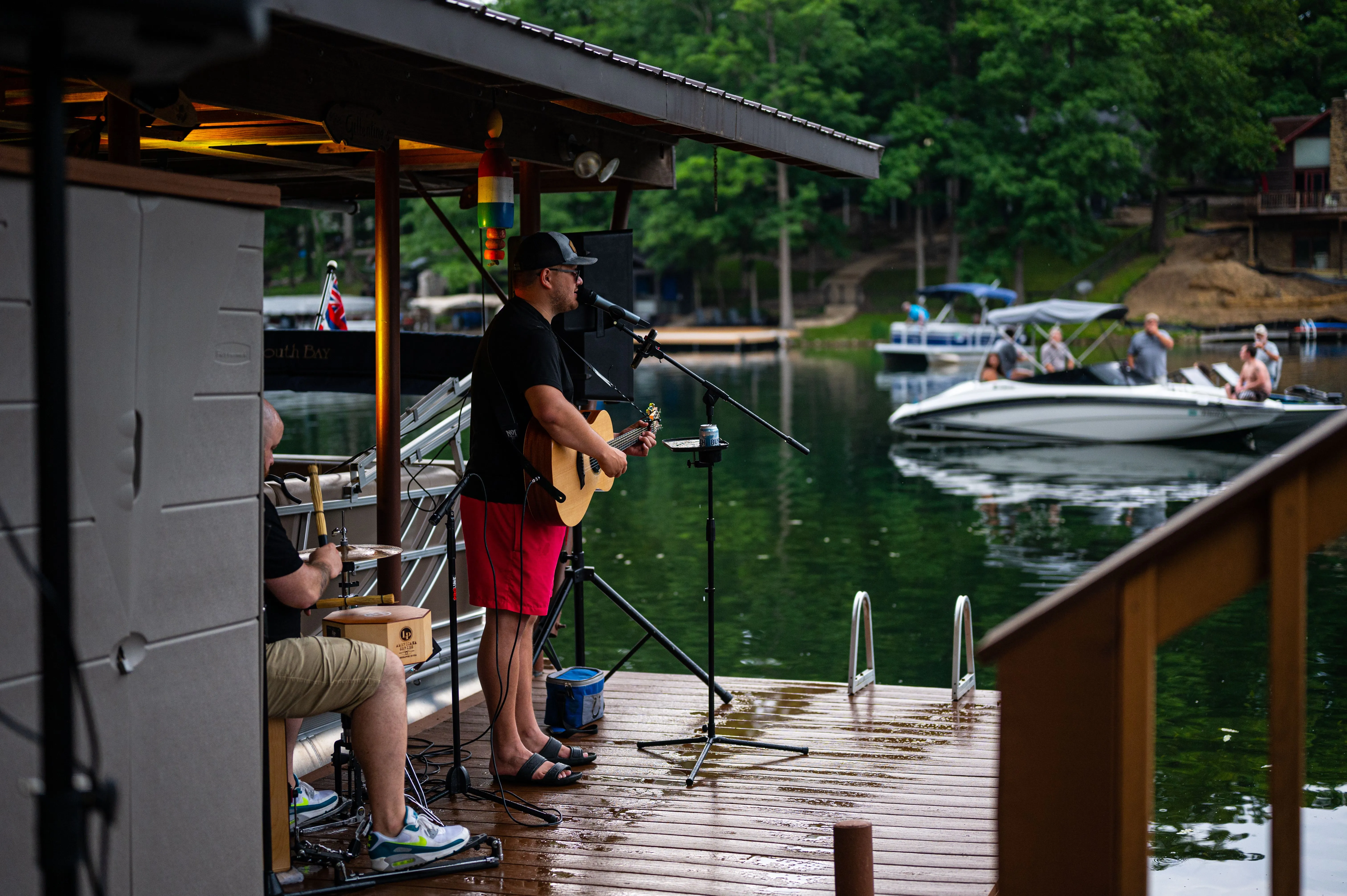 Two musicians performing on a lakeside dock with boats in the background.