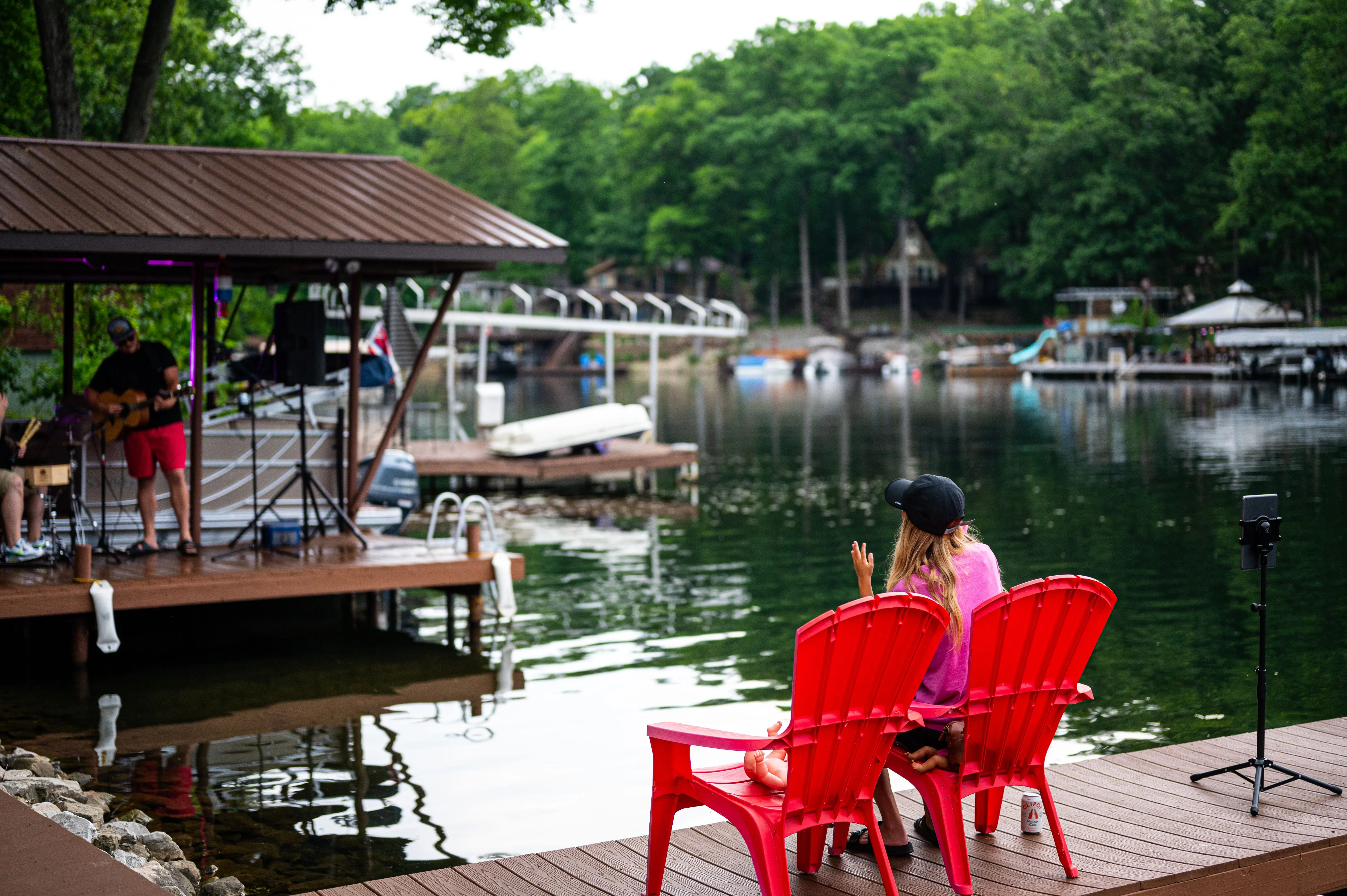  A person sitting on a red chair by a calm lake, gazing out at a dock with boats and a gazebo with a musician performing.