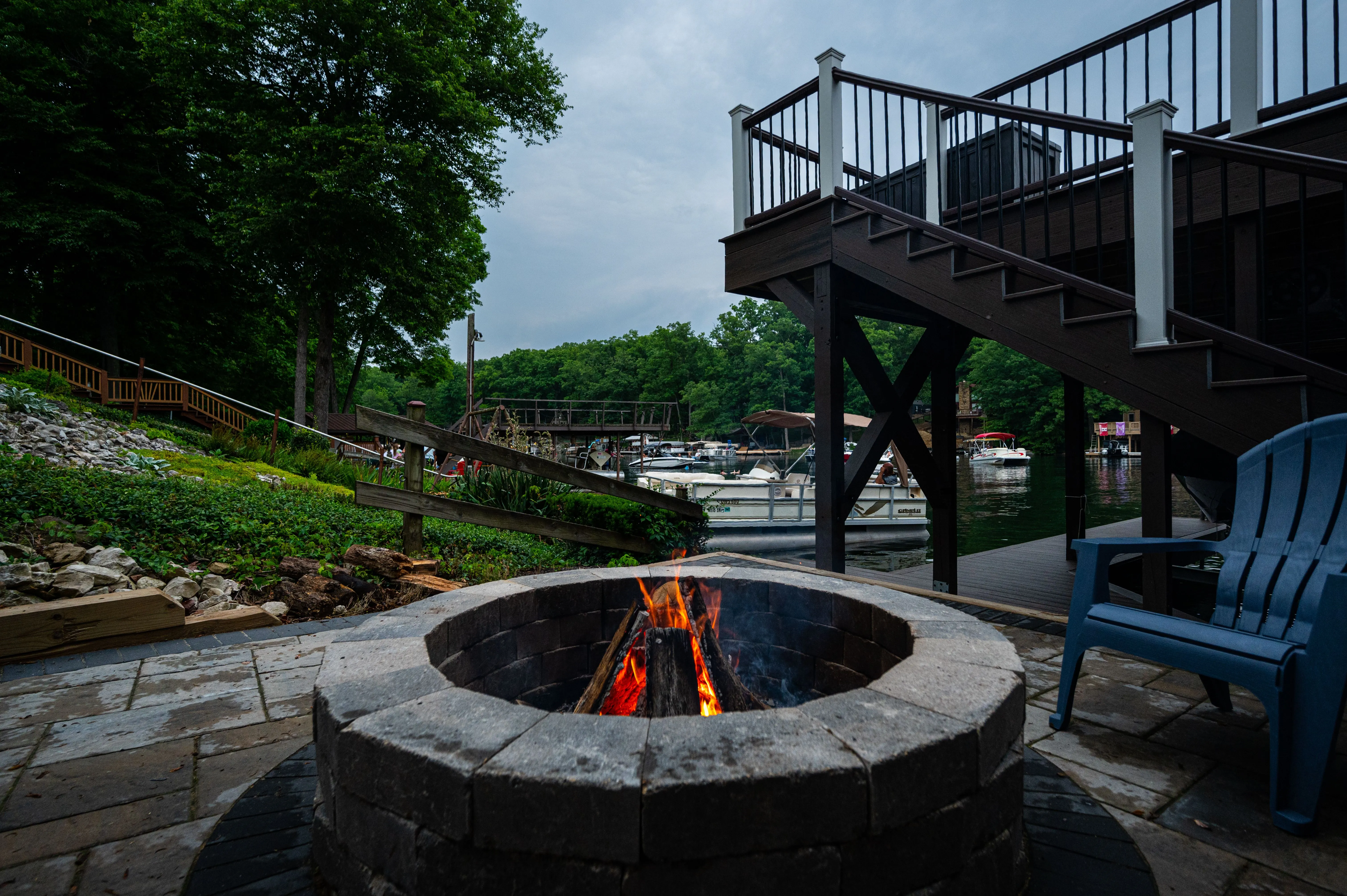 An outdoor stone fire pit with a lit fire, surrounded by seating, with stairs and lush greenery in the background.