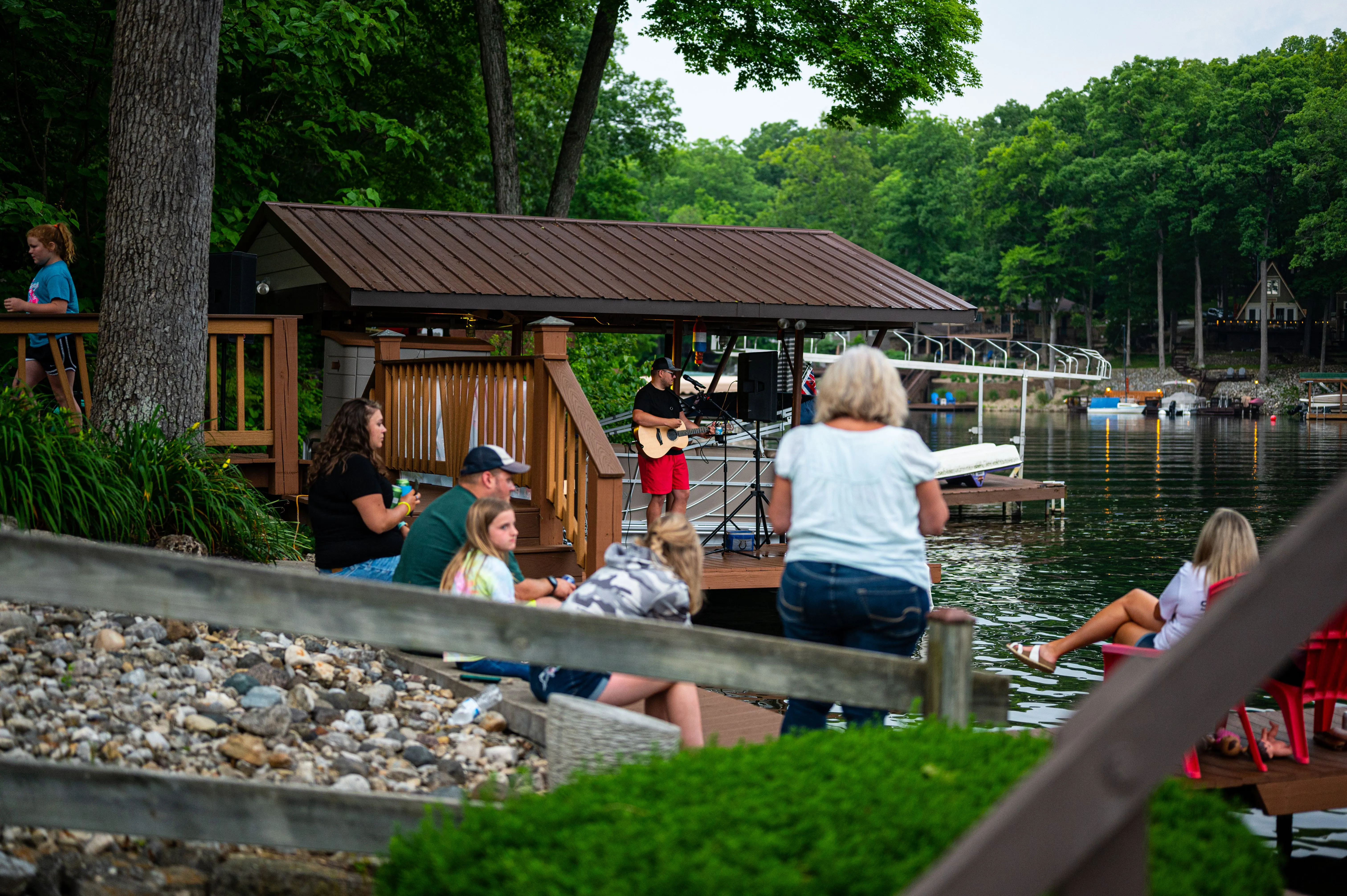 People relaxing by a lakeside with a small wooden cabin and dock in the background.
