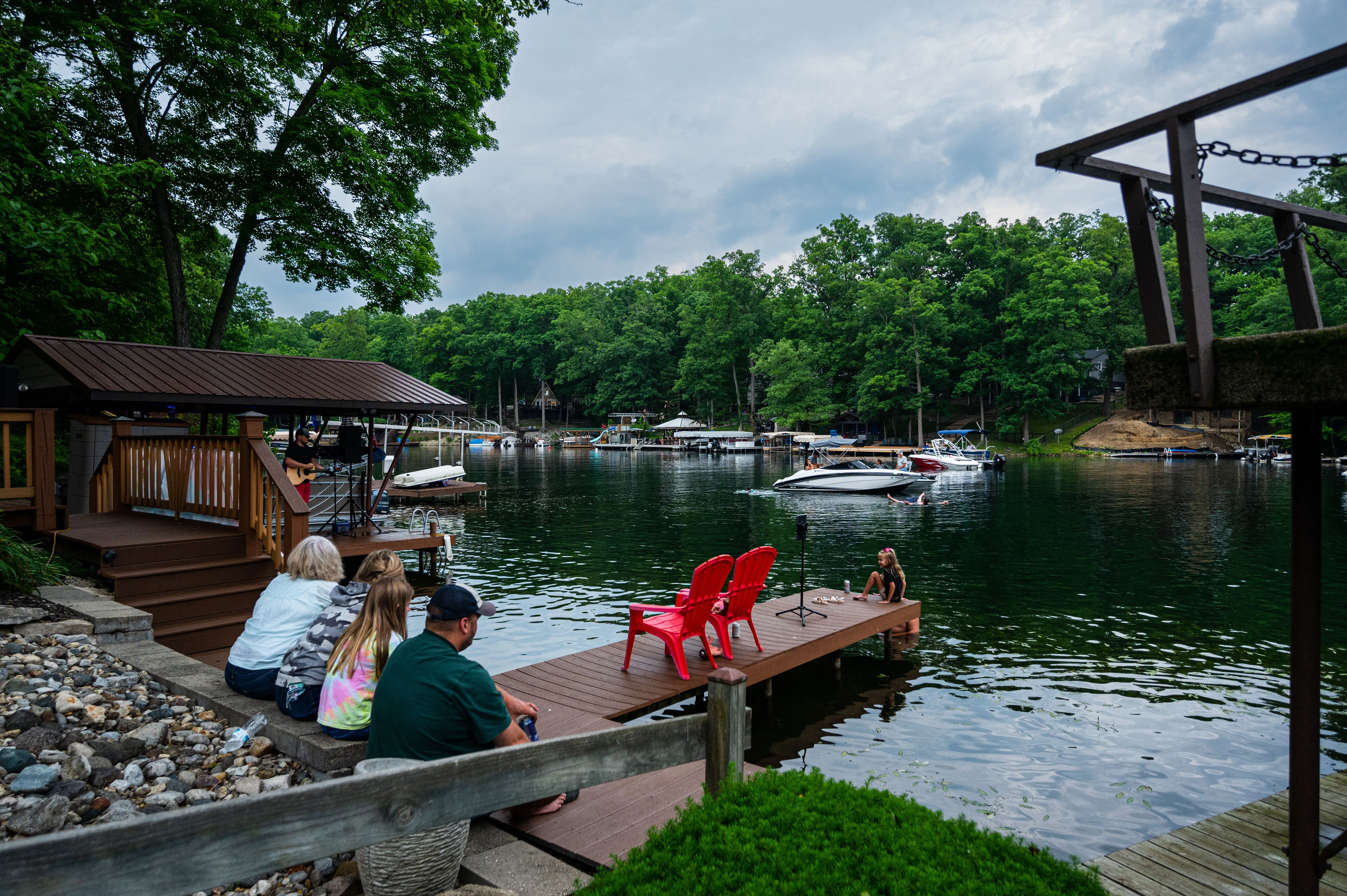 People relaxing by a calm lake with a dock, boats, and forested shores, capturing a serene atmosphere.