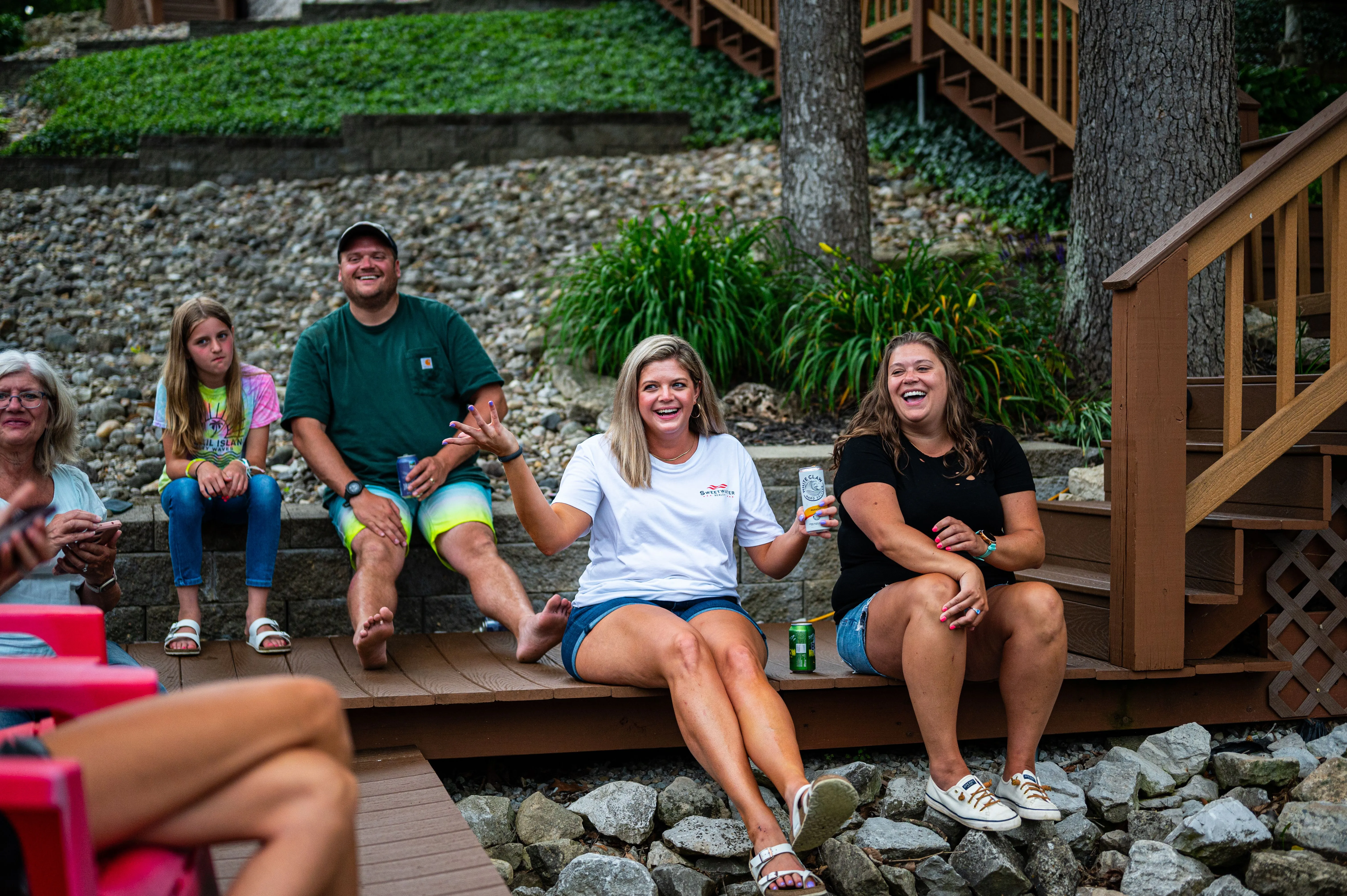 Group of people smiling and sitting on a wooden dock, with one person holding a green plastic cup, in a backyard setting with stairs and gravel.