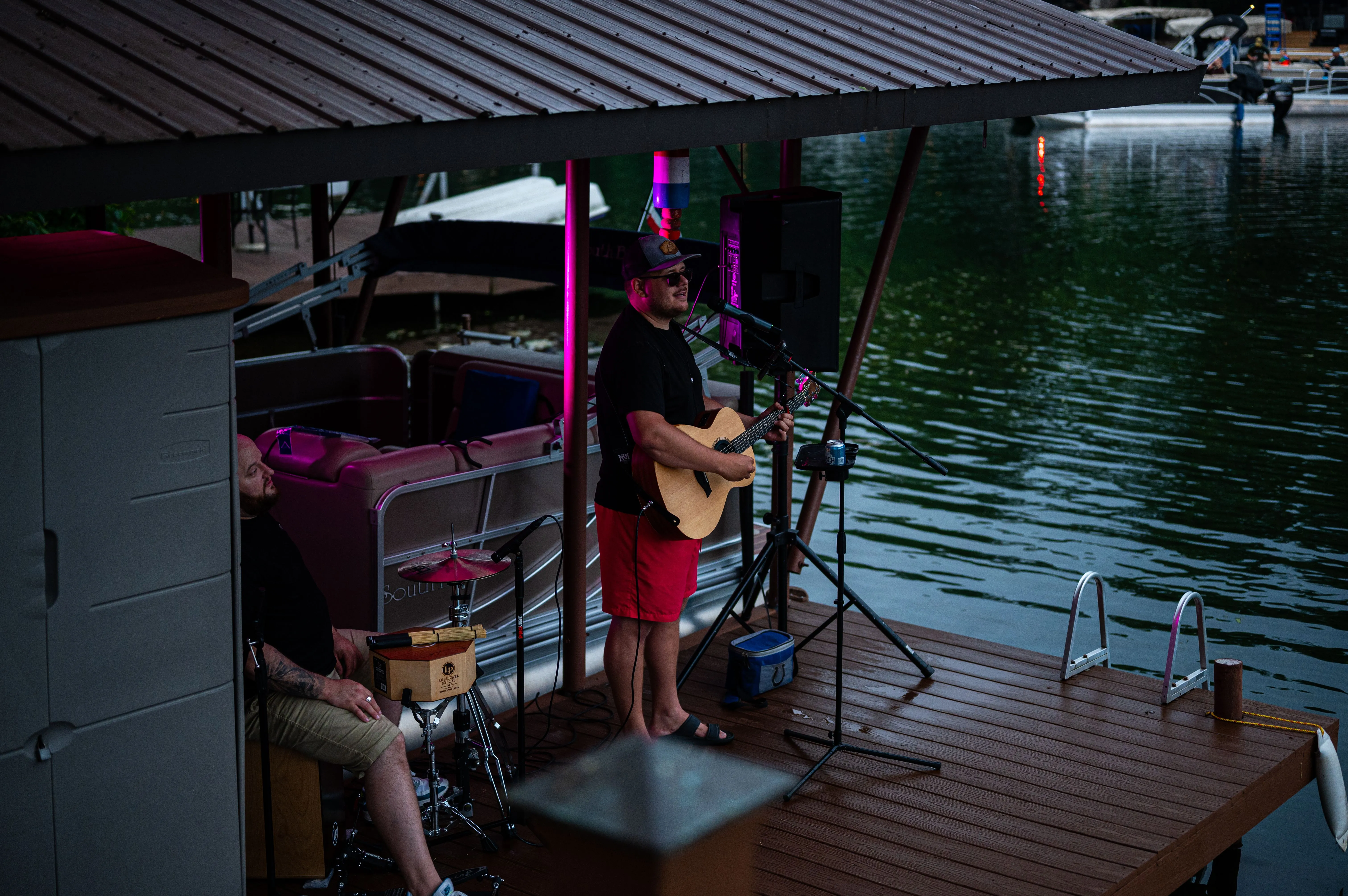 Musician playing guitar on a floating dock stage under a shelter at dusk.
