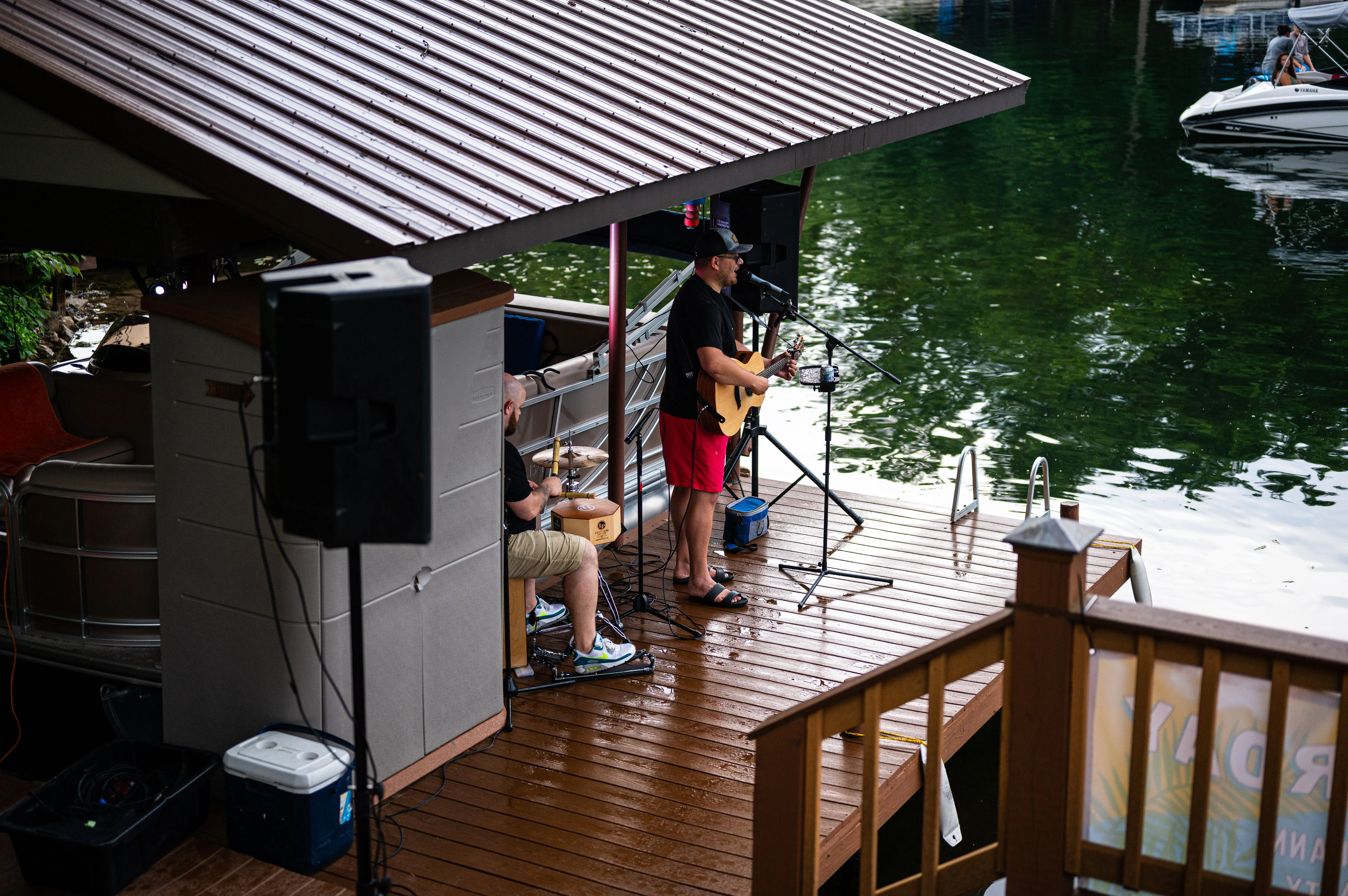 Man playing guitar on a covered dock by the water with a dog sitting nearby.