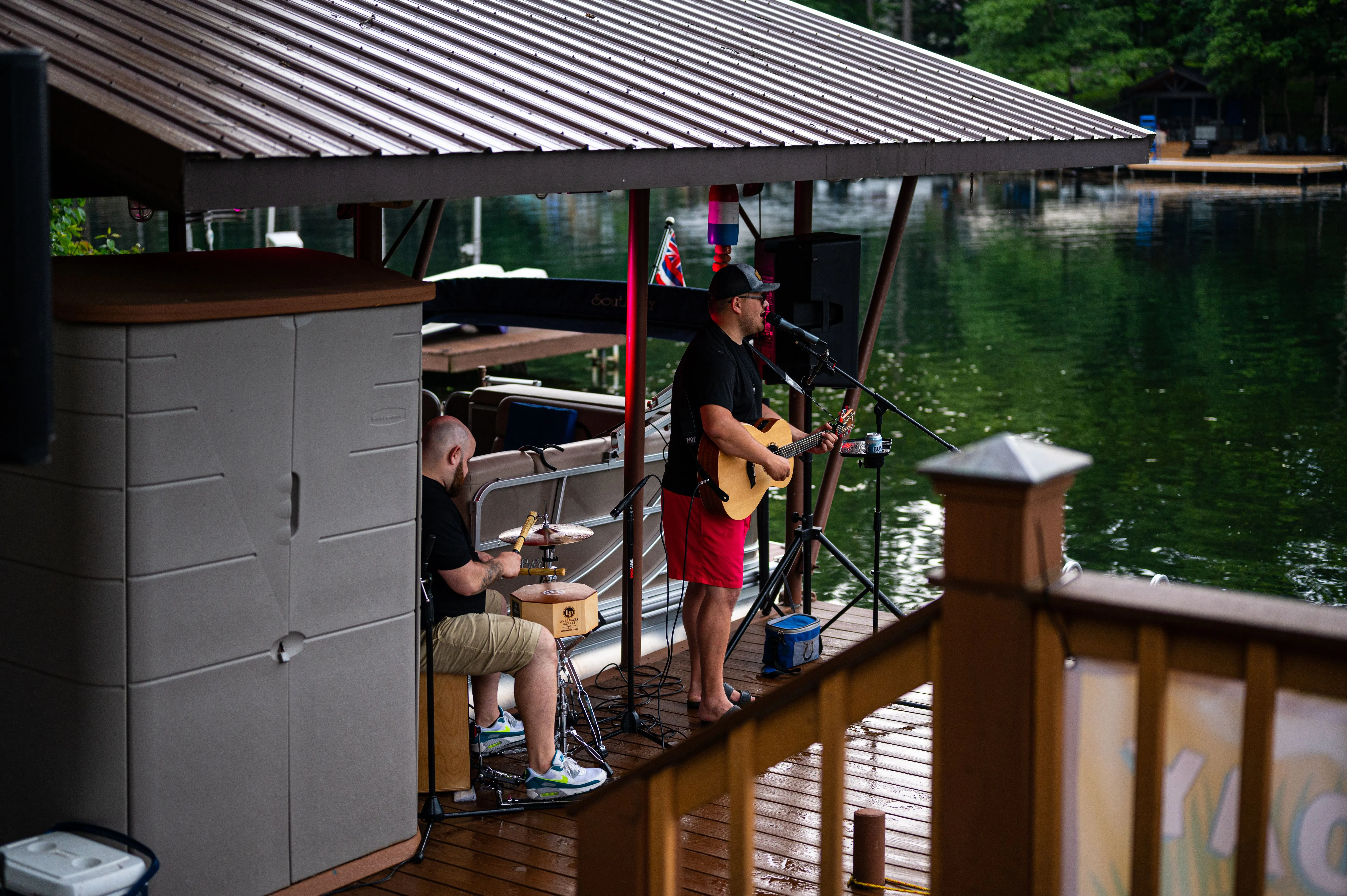 Two musicians performing on a covered dock by a lake, one playing drums and the other playing guitar.