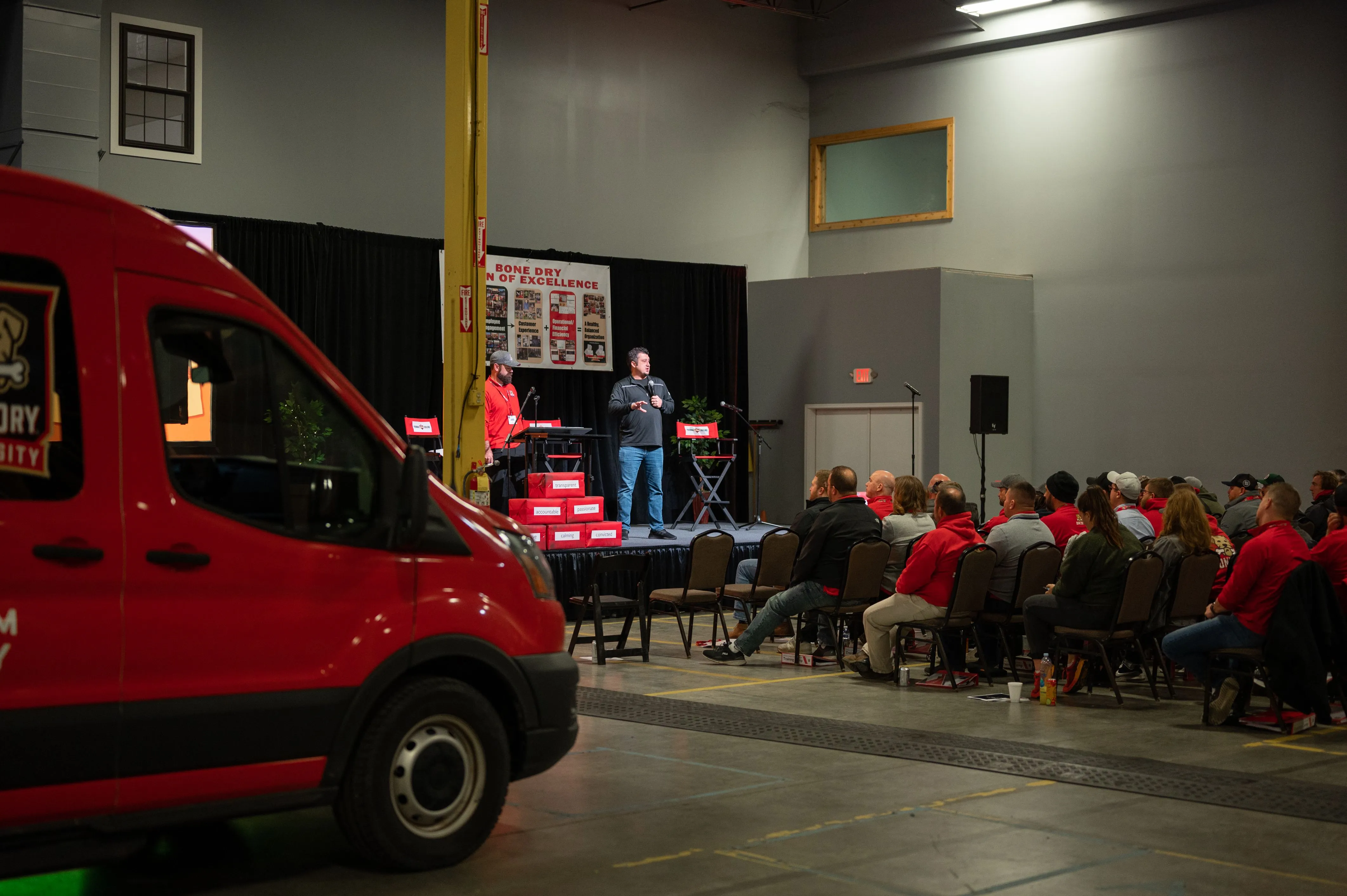 Red van parked indoors with a person speaking on a stage to an audience in the background.