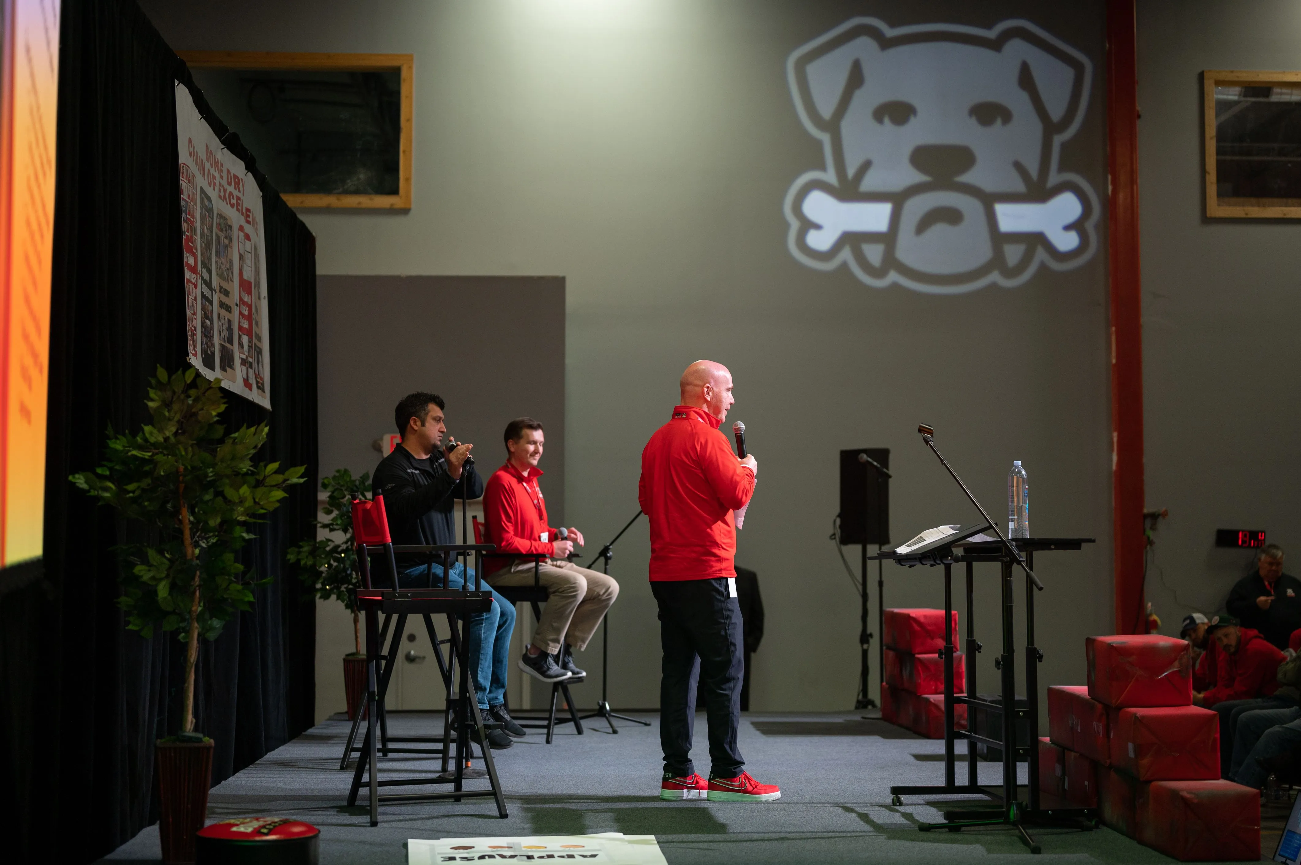 Three people on stage at an event with a cartoon dog logo projected on the wall behind them.