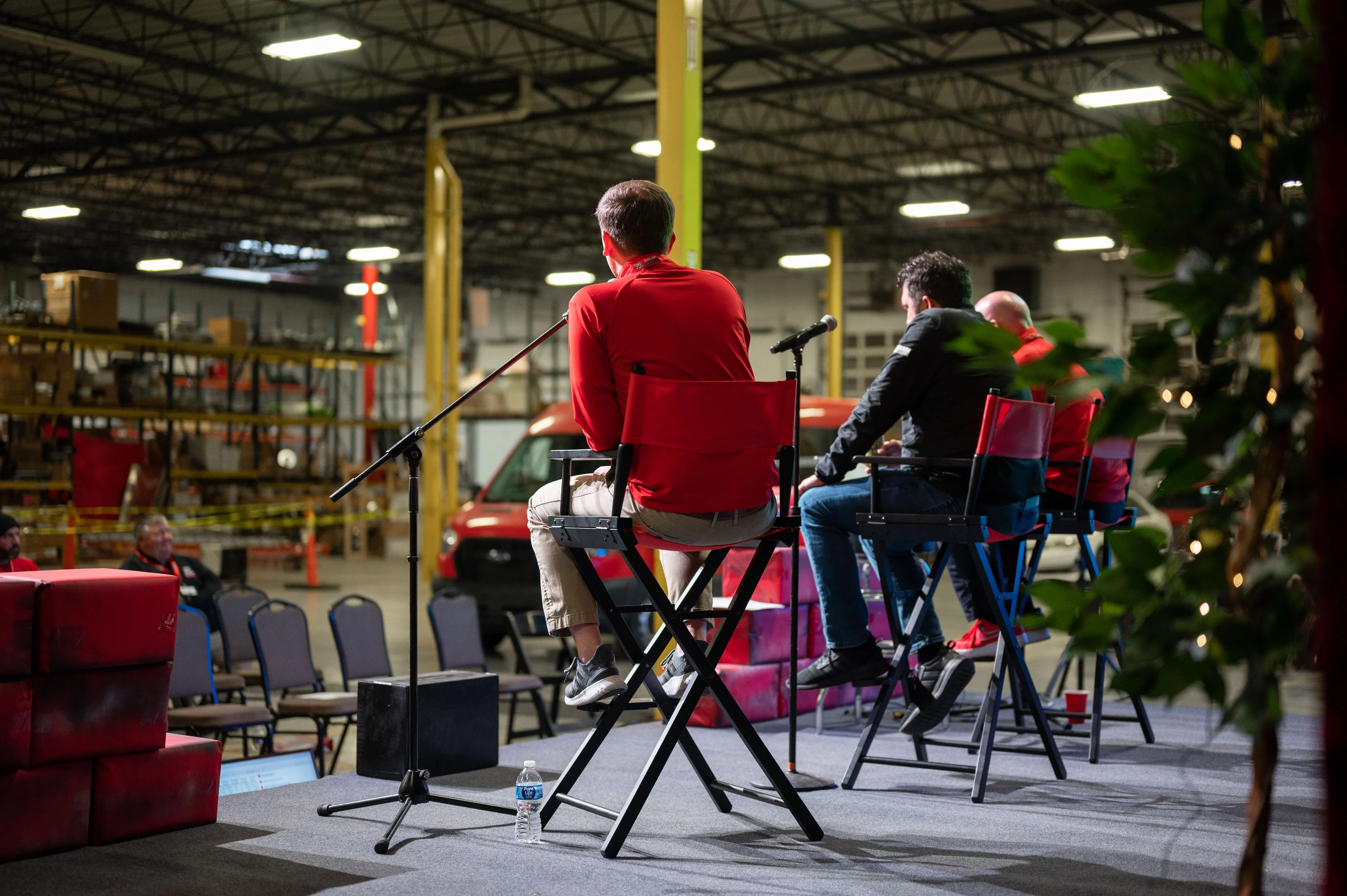 Three people sitting on high chairs in an industrial setting, possibly engaged in a discussion panel or interview, with warehouse shelves in the background.