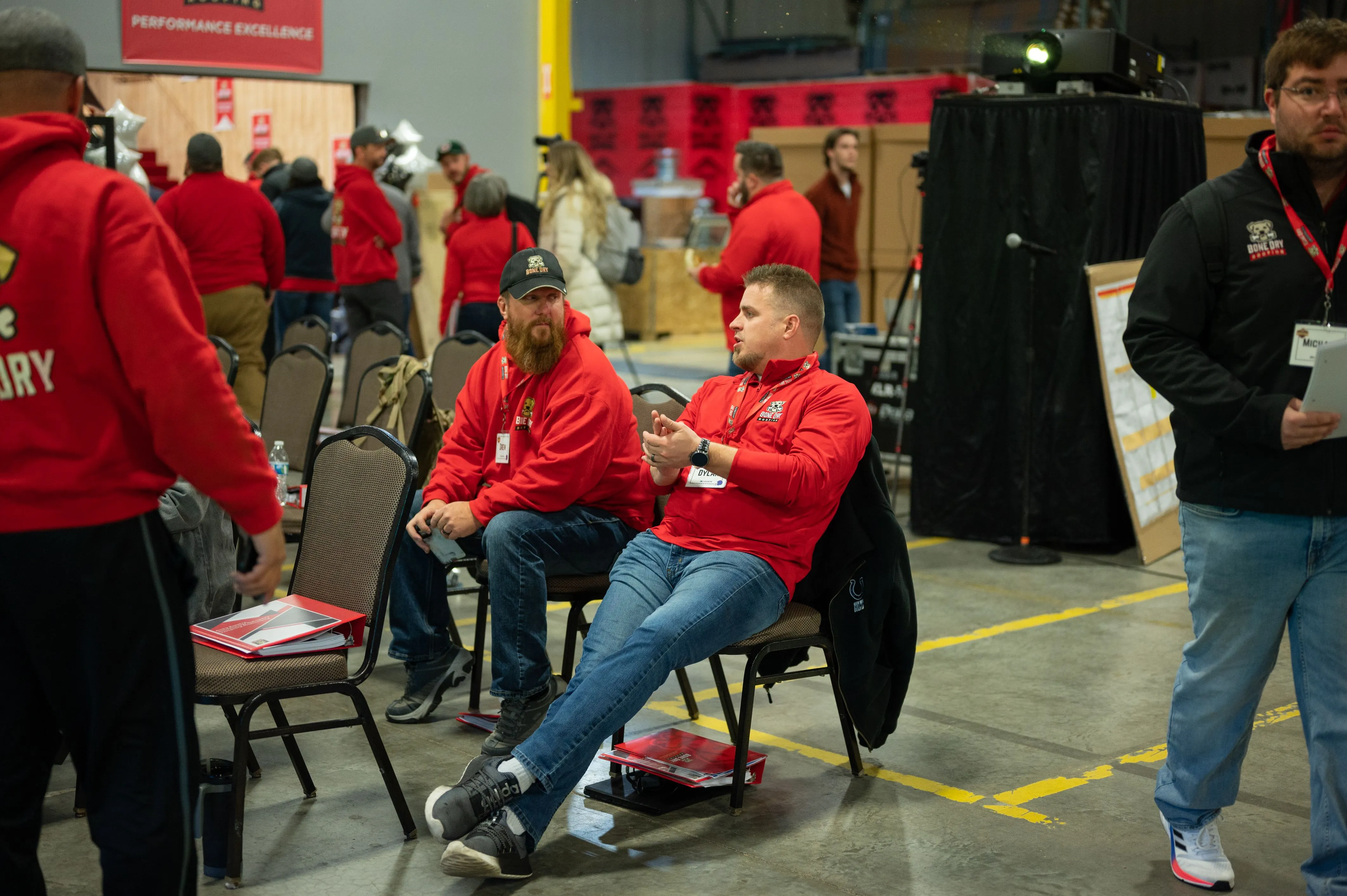 People wearing red tops at a busy indoor event with some sitting and one person using a mobile phone.