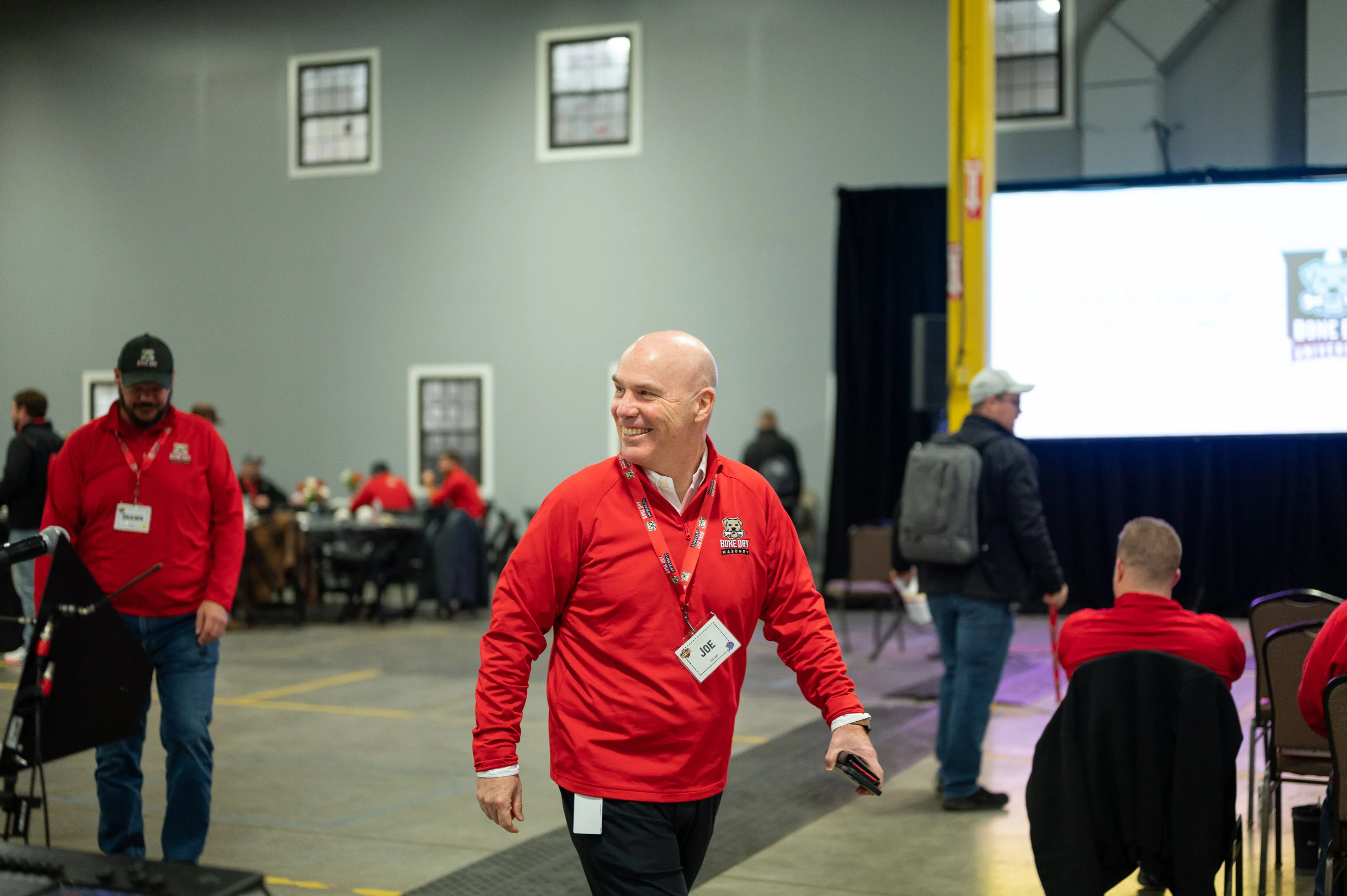 A man in a red jacket walking through a convention center with other attendees in the background.