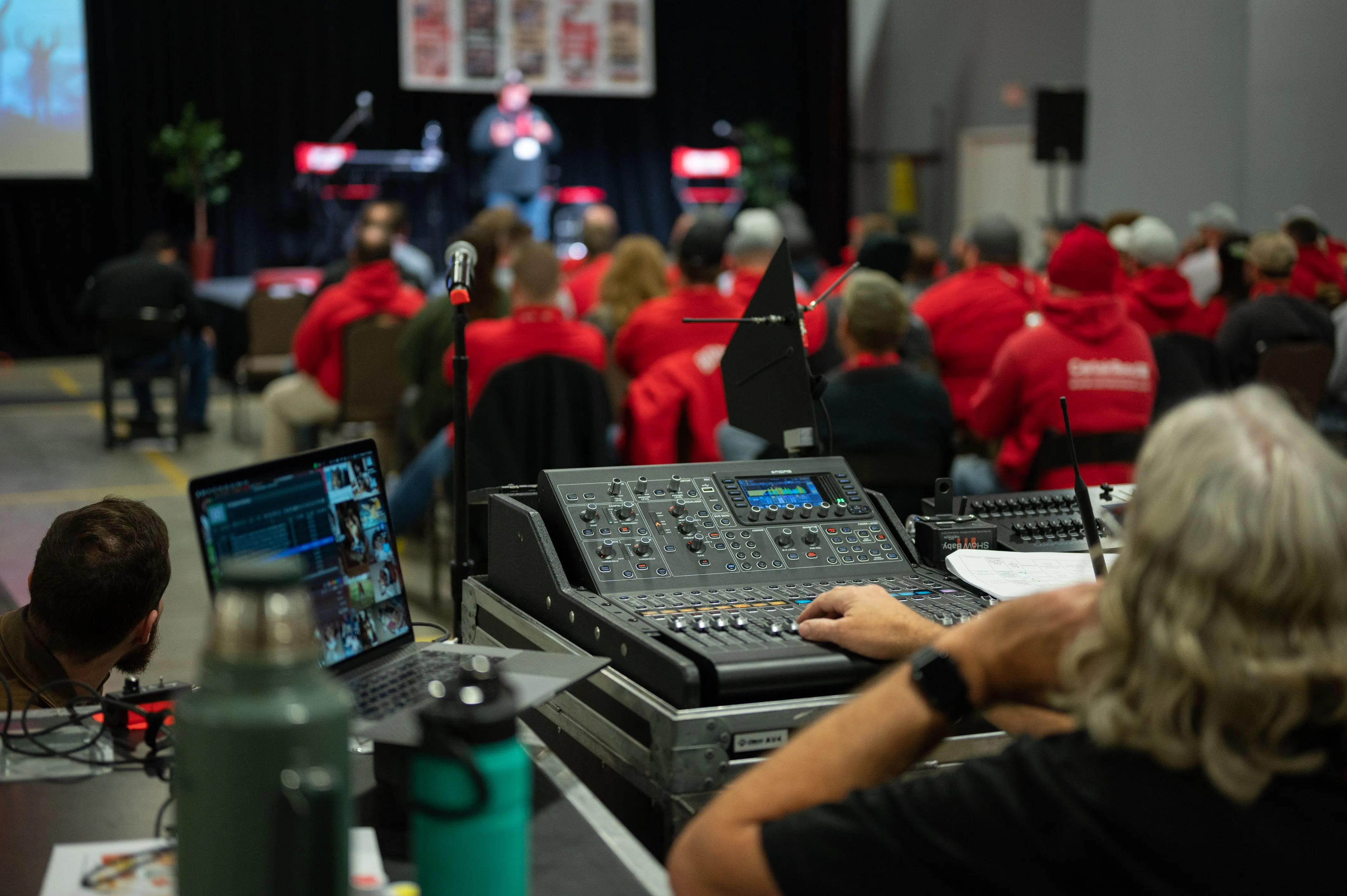 Audience in red shirts at a conference with a presenter in the background, foreground shows person operating an audio mixing console.