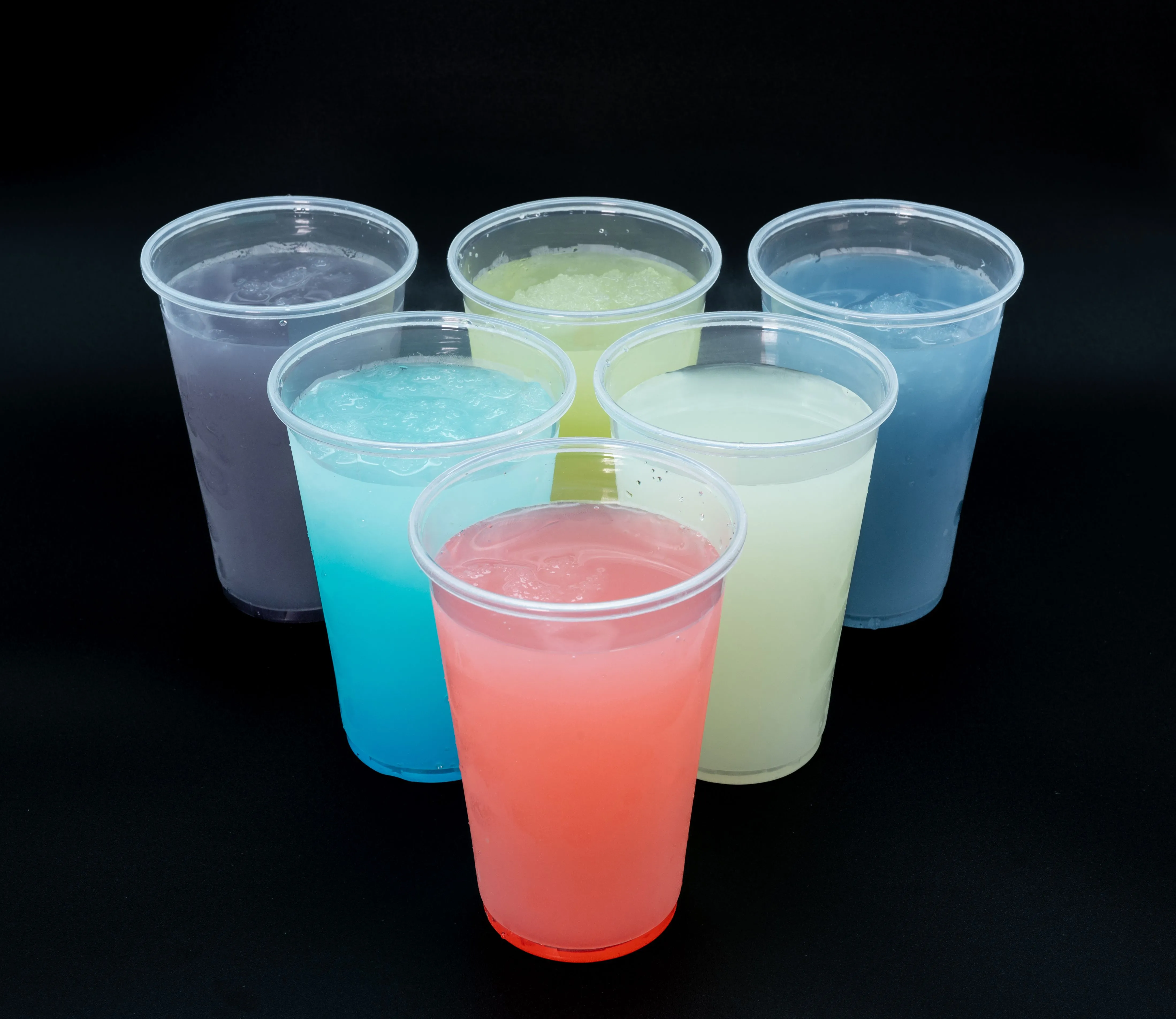 Seven colorful drinks in clear plastic cups on a black background.