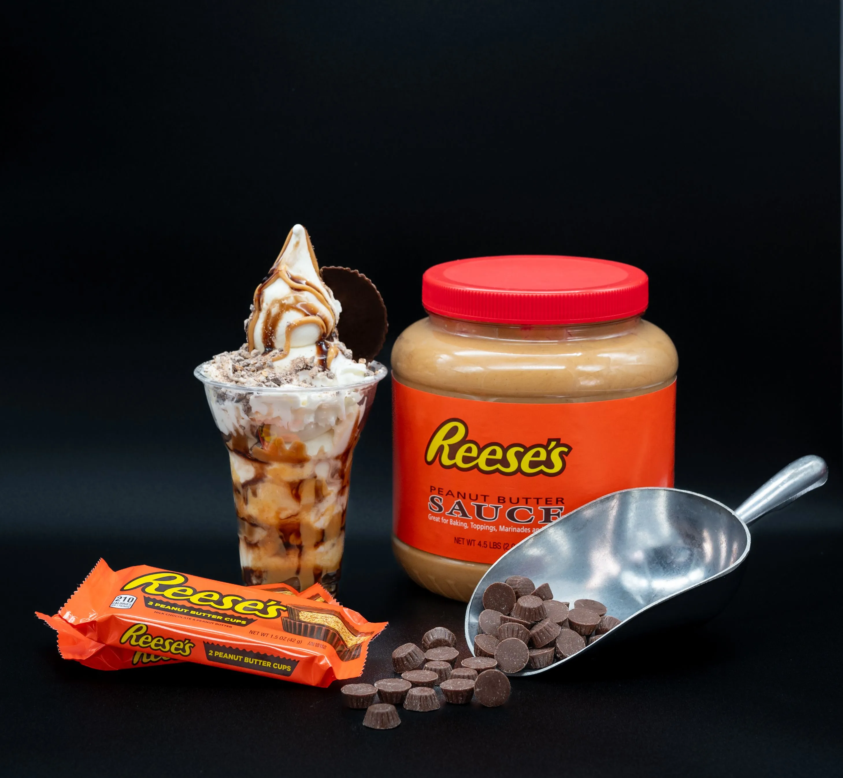 Reese's branded products including a peanut butter cup, peanut butter sauce jar, sundae with whipped cream, chocolate syrup, and a chocolate cookie, chocolate chips, and a metal scooping spoon against a black background.