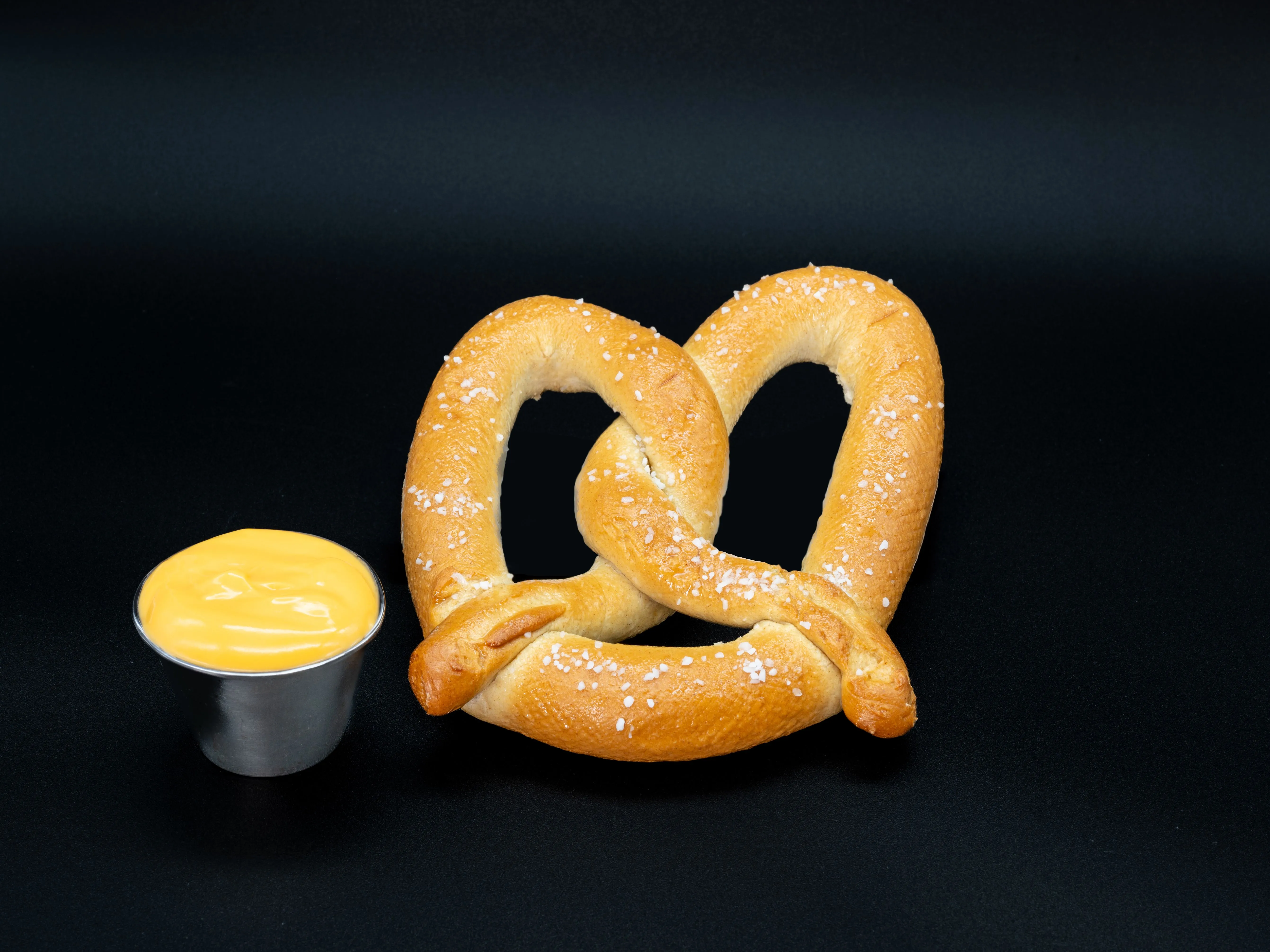 A baked pretzel with salt crystals on top alongside a small container of cheese sauce, both placed on a black background.
