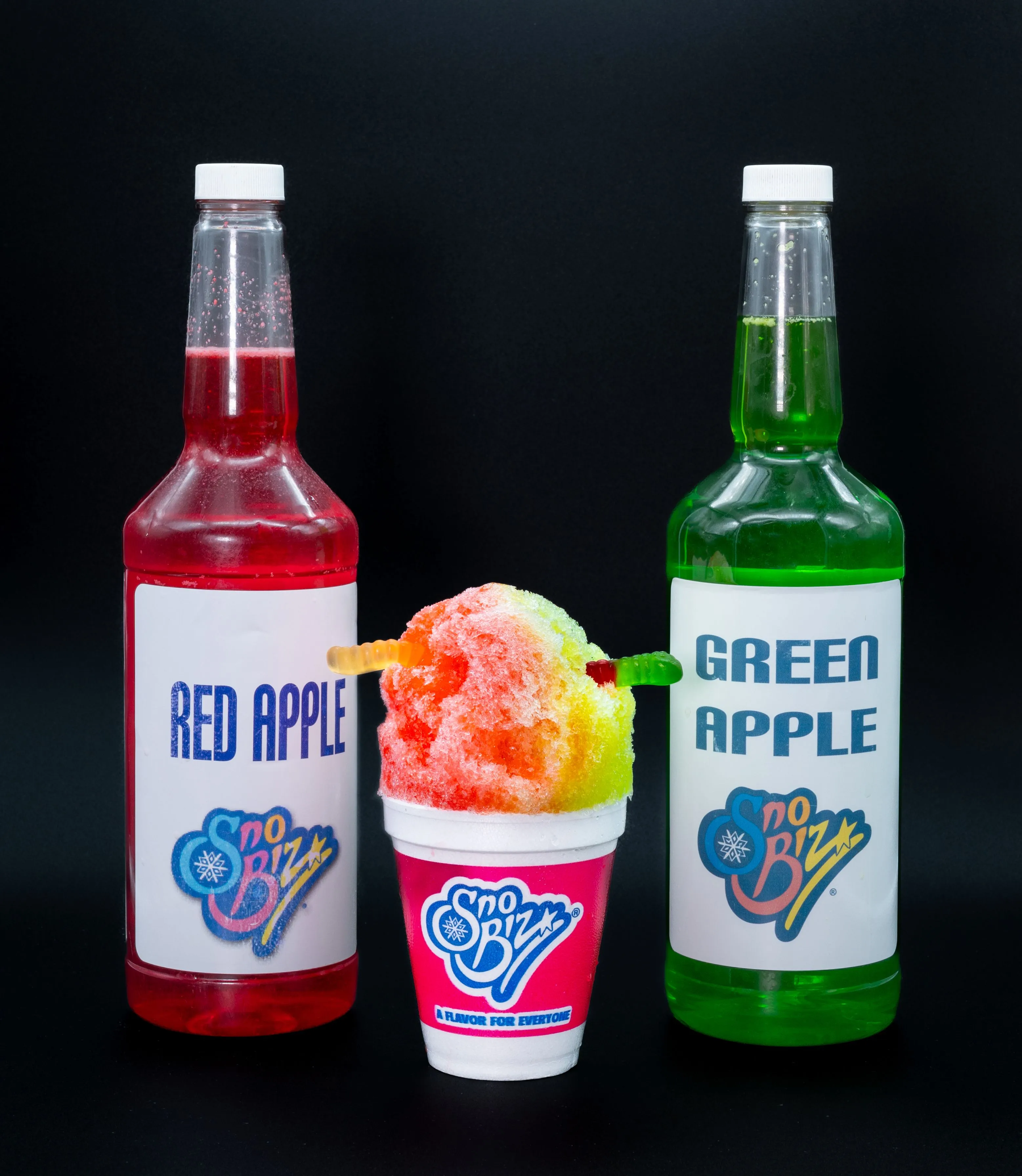 Two bottles labeled 'Red Apple' and 'Green Apple' flanking a colorful shaved ice in a Sno Biz cup on a black background.