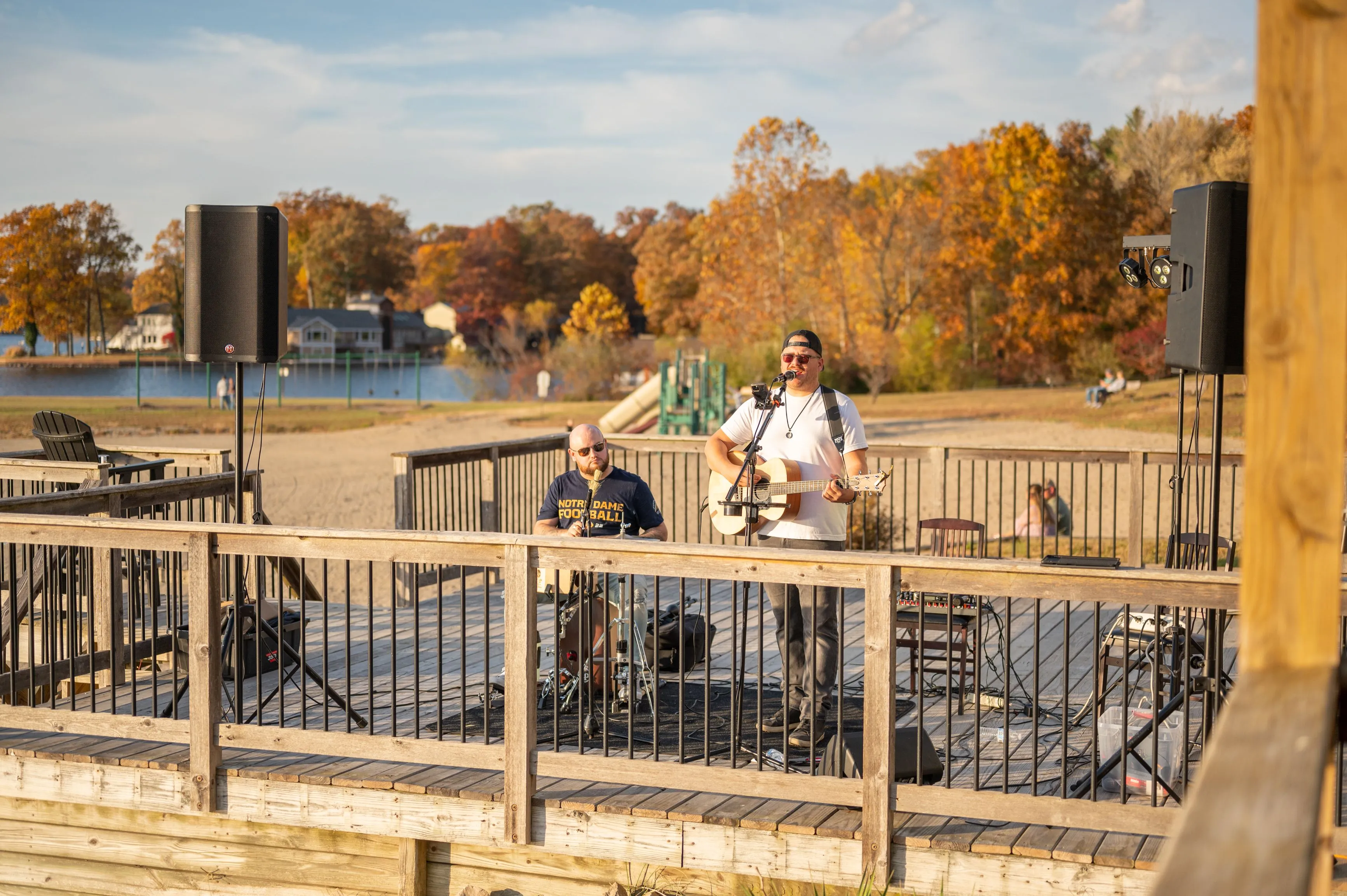Two musicians performing outside on a waterfront deck with empty chairs and autumn trees in the background.