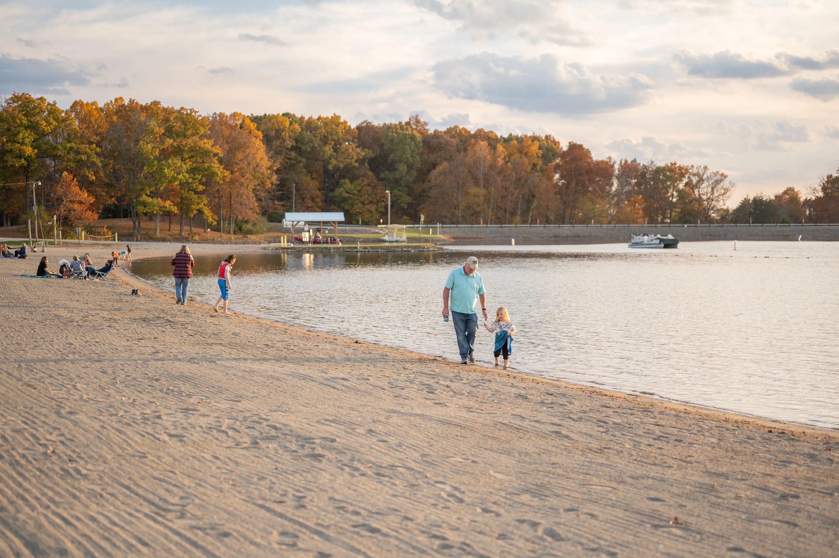 An adult and a child walking hand in hand on a sandy beach with other visitors and autumn trees in the background.