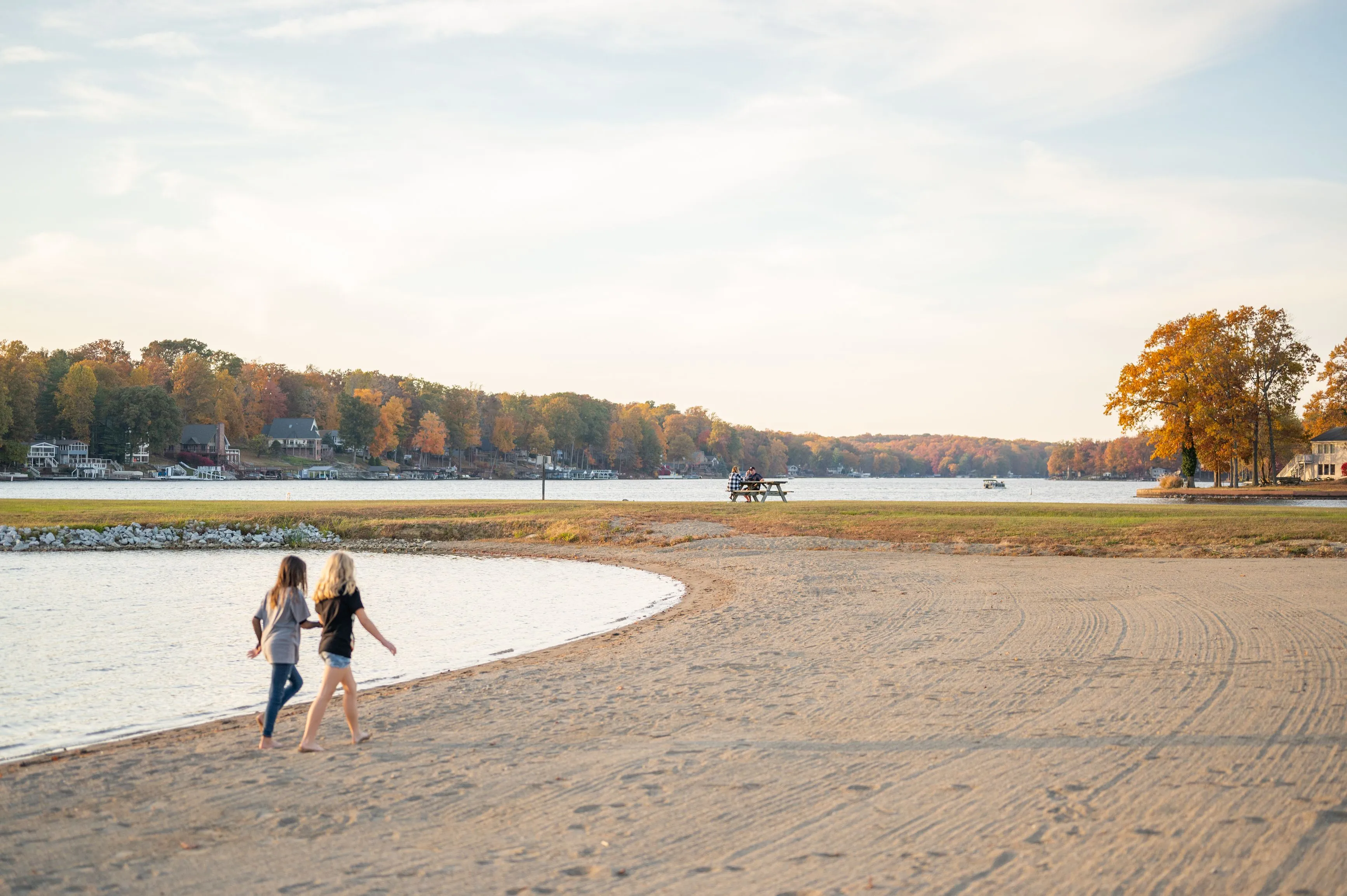 Two individuals walking on a sandy beach with trees and a lake in the background during autumn.