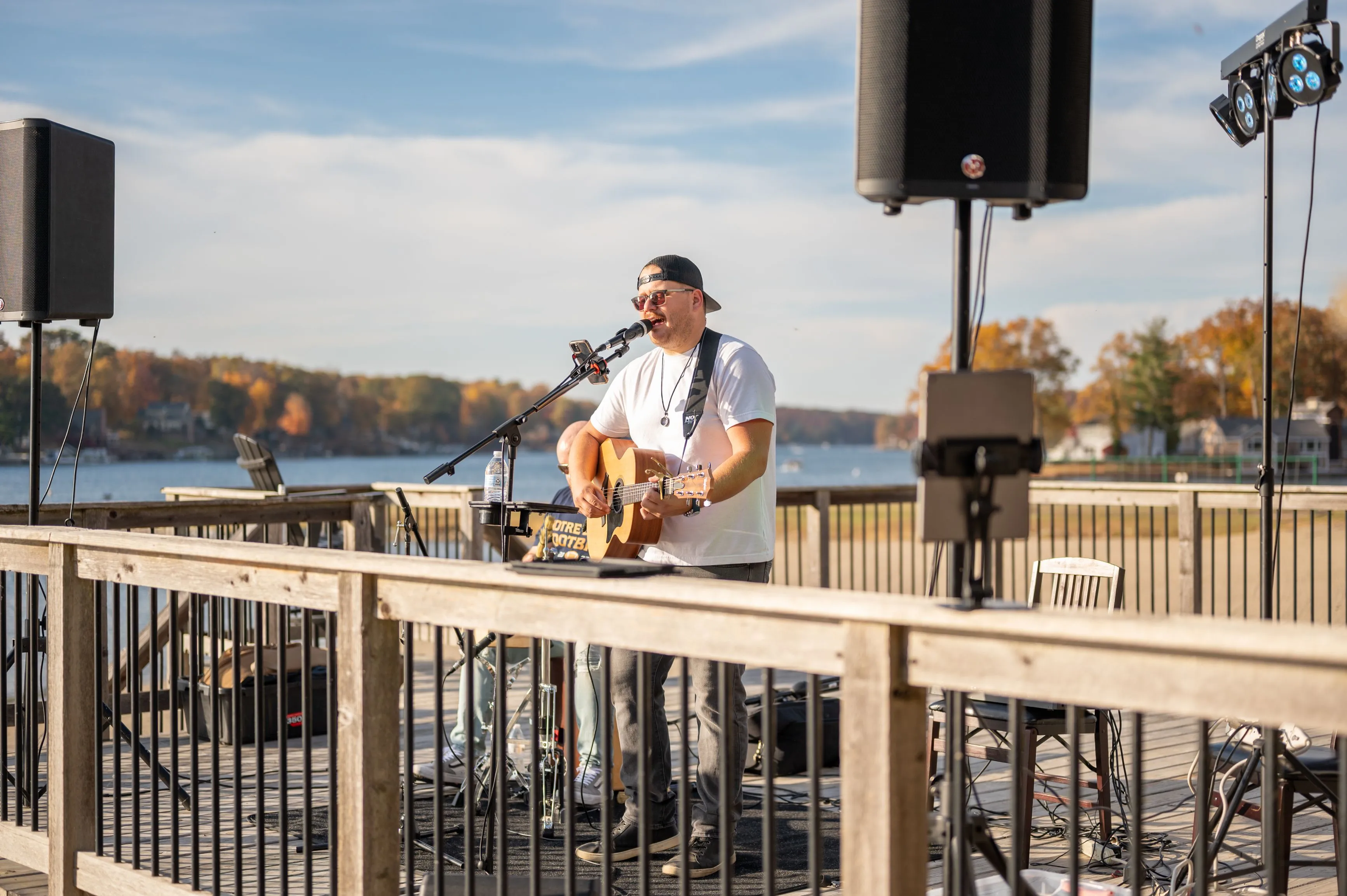 Musician playing guitar and singing into a microphone on an outdoor stage by a lake.