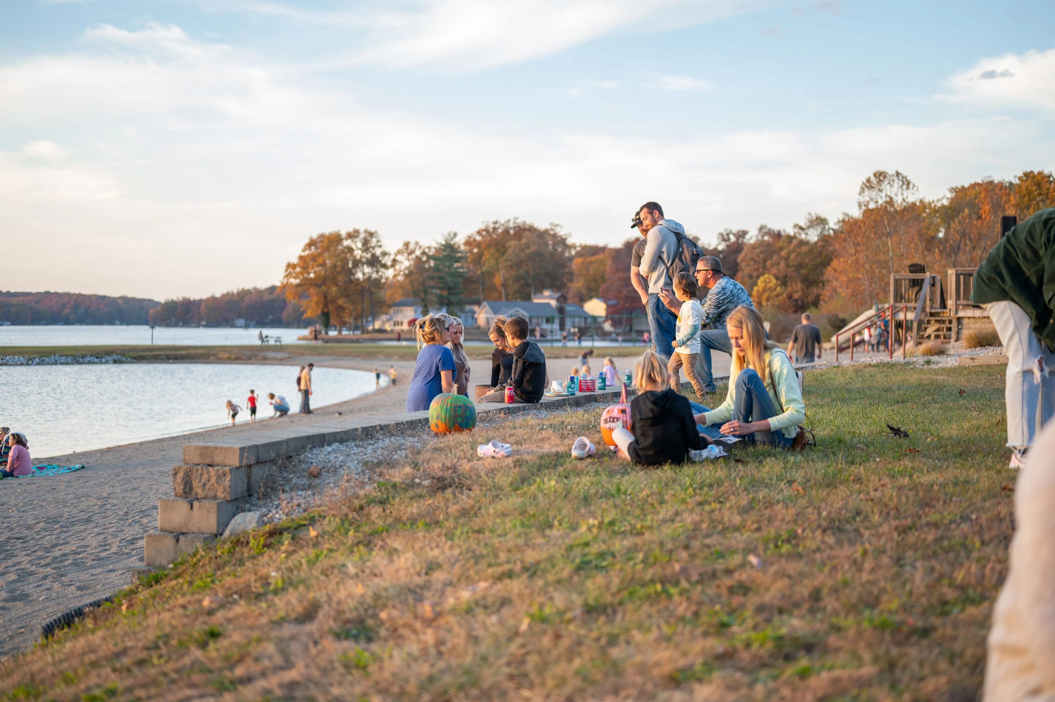People enjoying a sunny day at a lakeside park with some sitting on the grass and others standing.