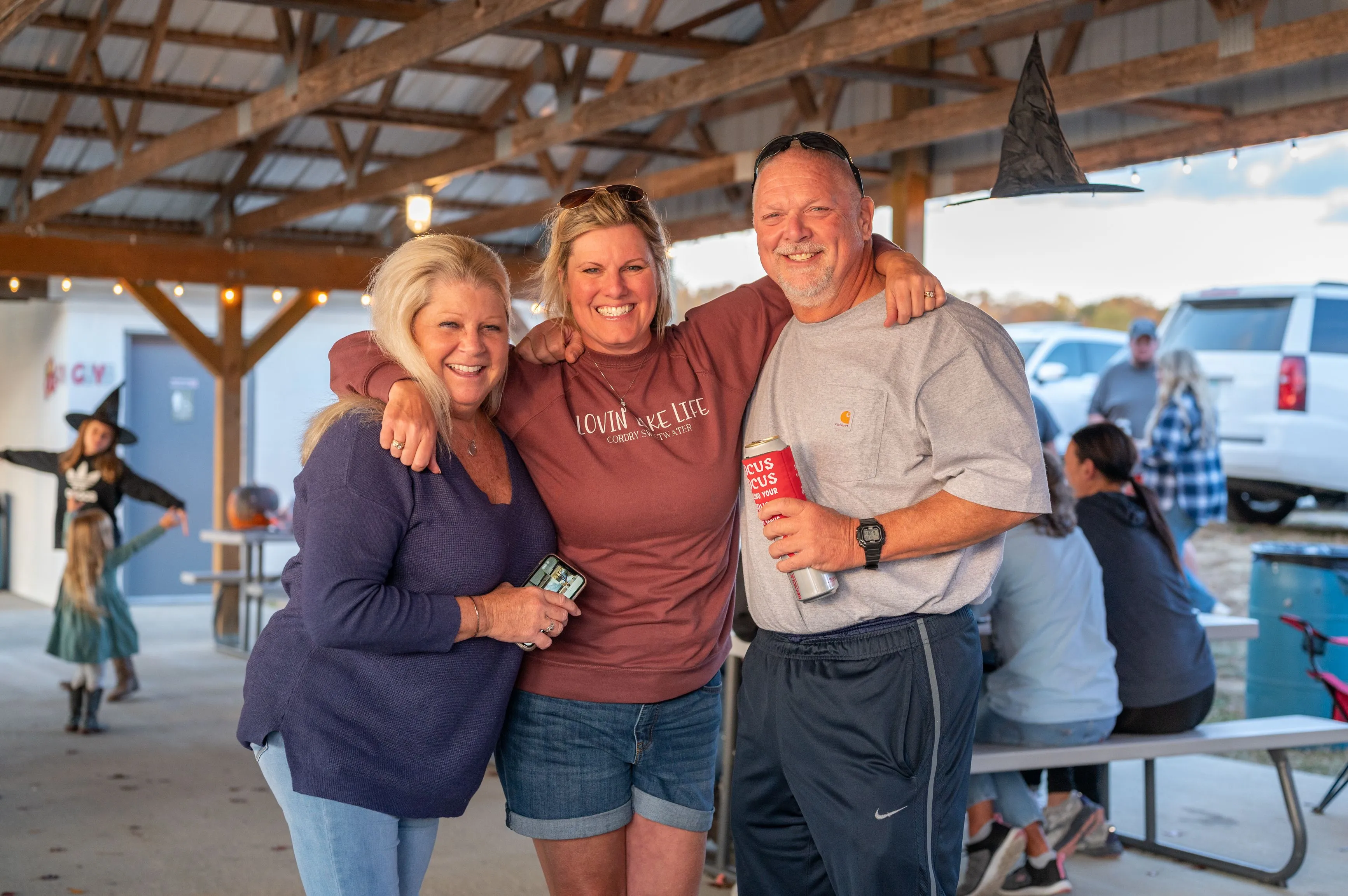 Three people smiling and posing for a photo at an outdoor gathering under a shelter.