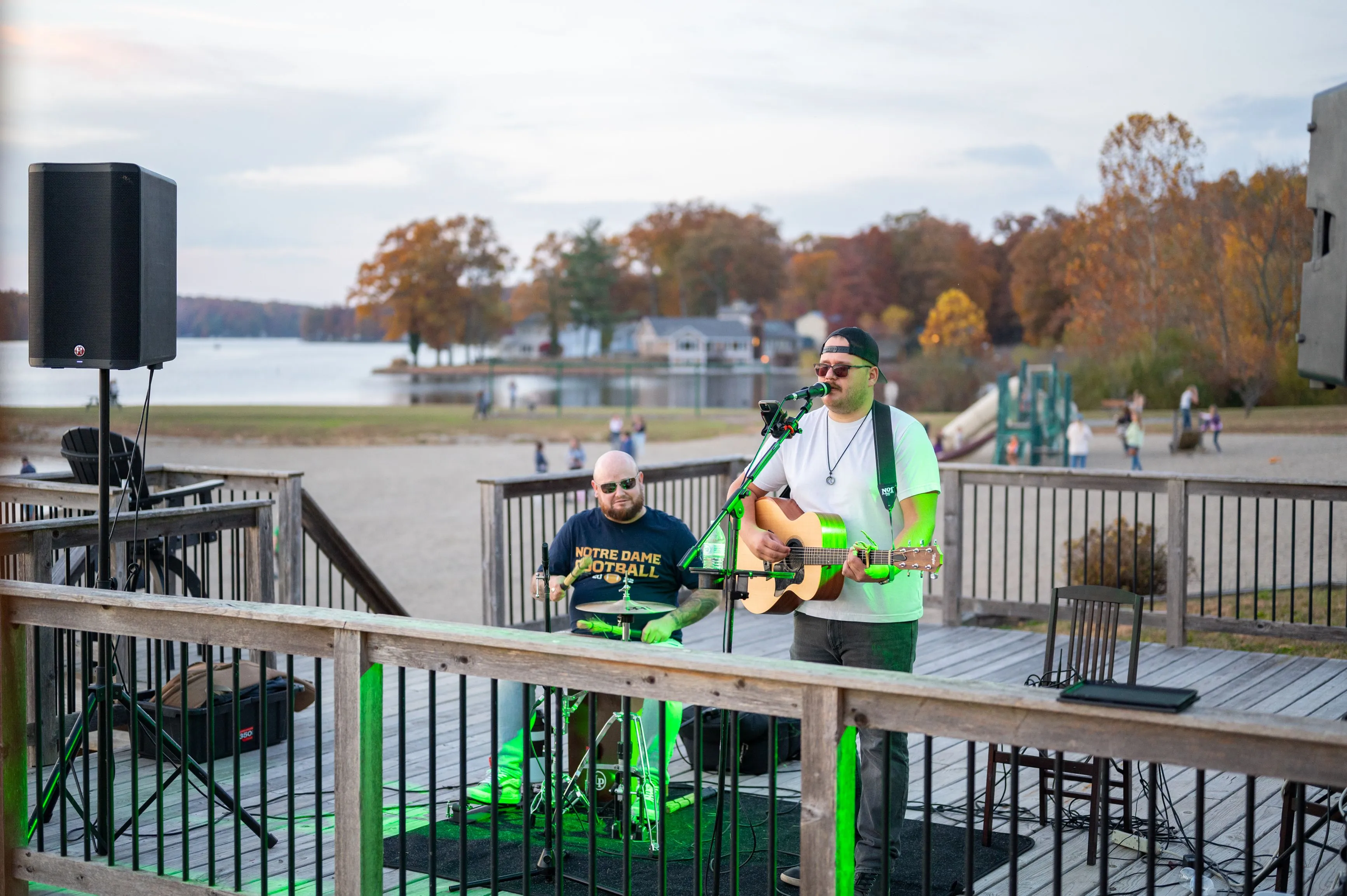 Two musicians performing outdoors on a deck overlooking a lake with autumn trees in the background.