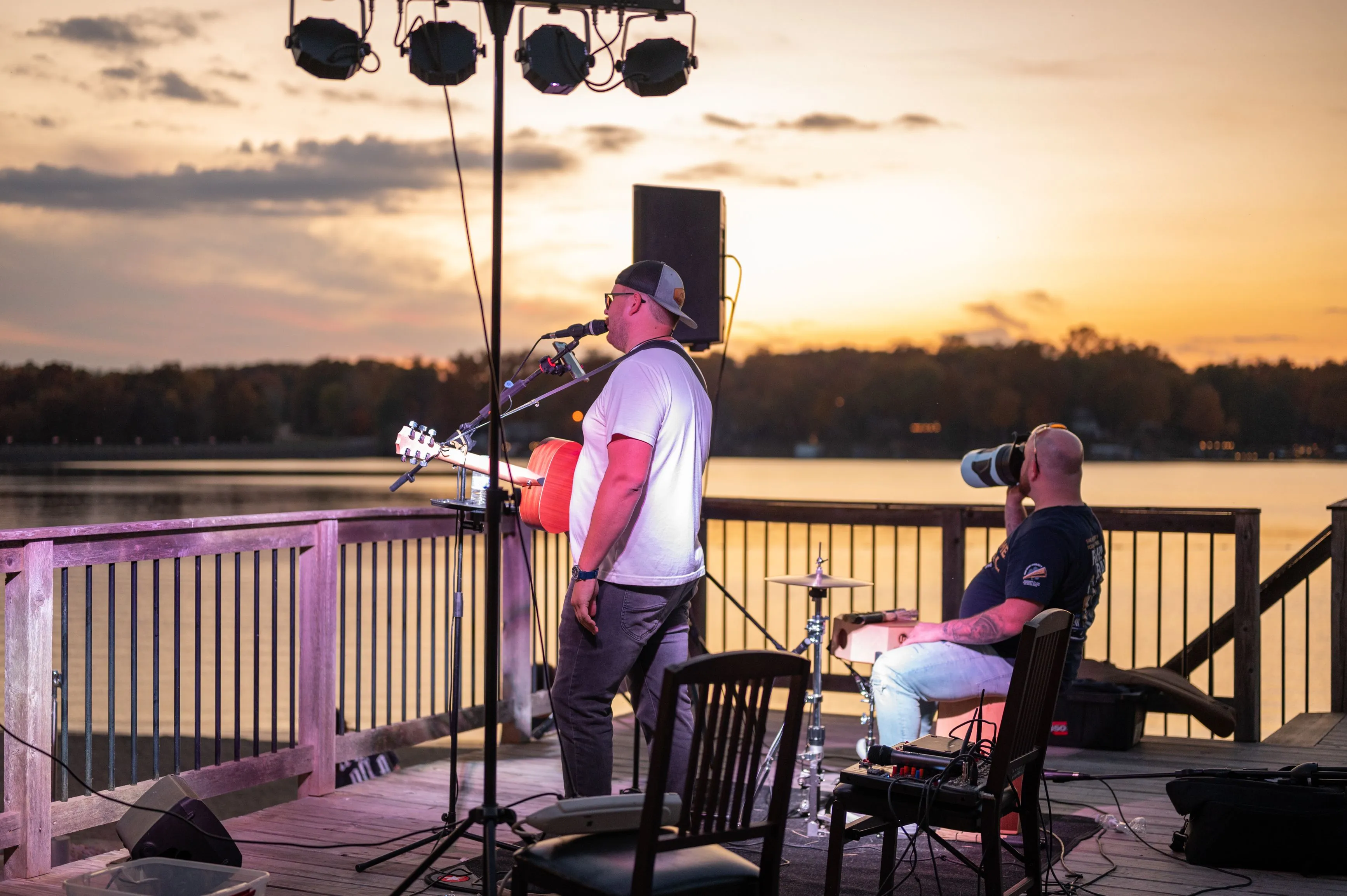 Musician playing guitar and singing into a microphone on an outdoor stage during sunset, with another person sitting by, near a body of water.