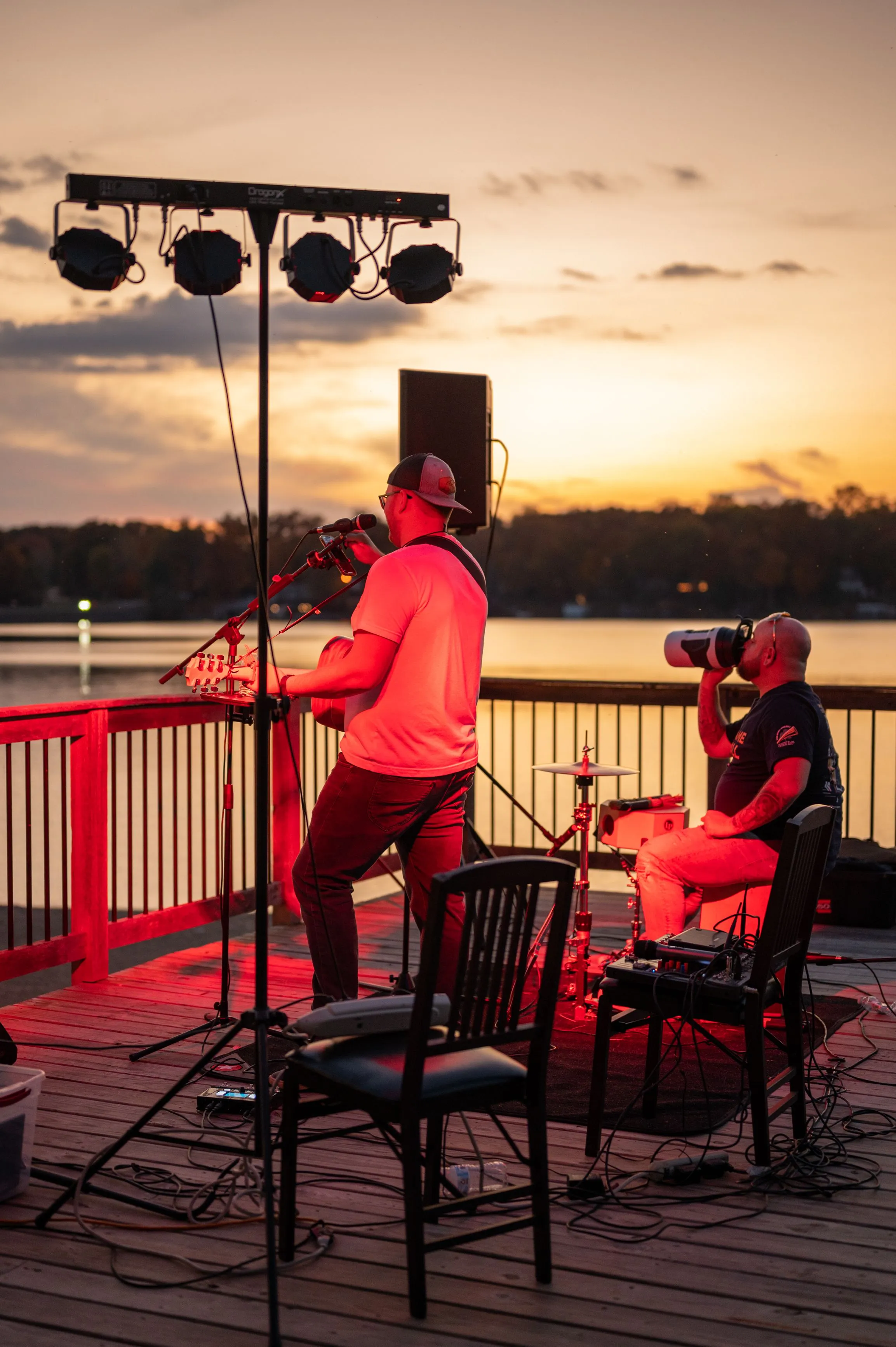 Musicians performing on an outdoor stage by a lake at sunset.