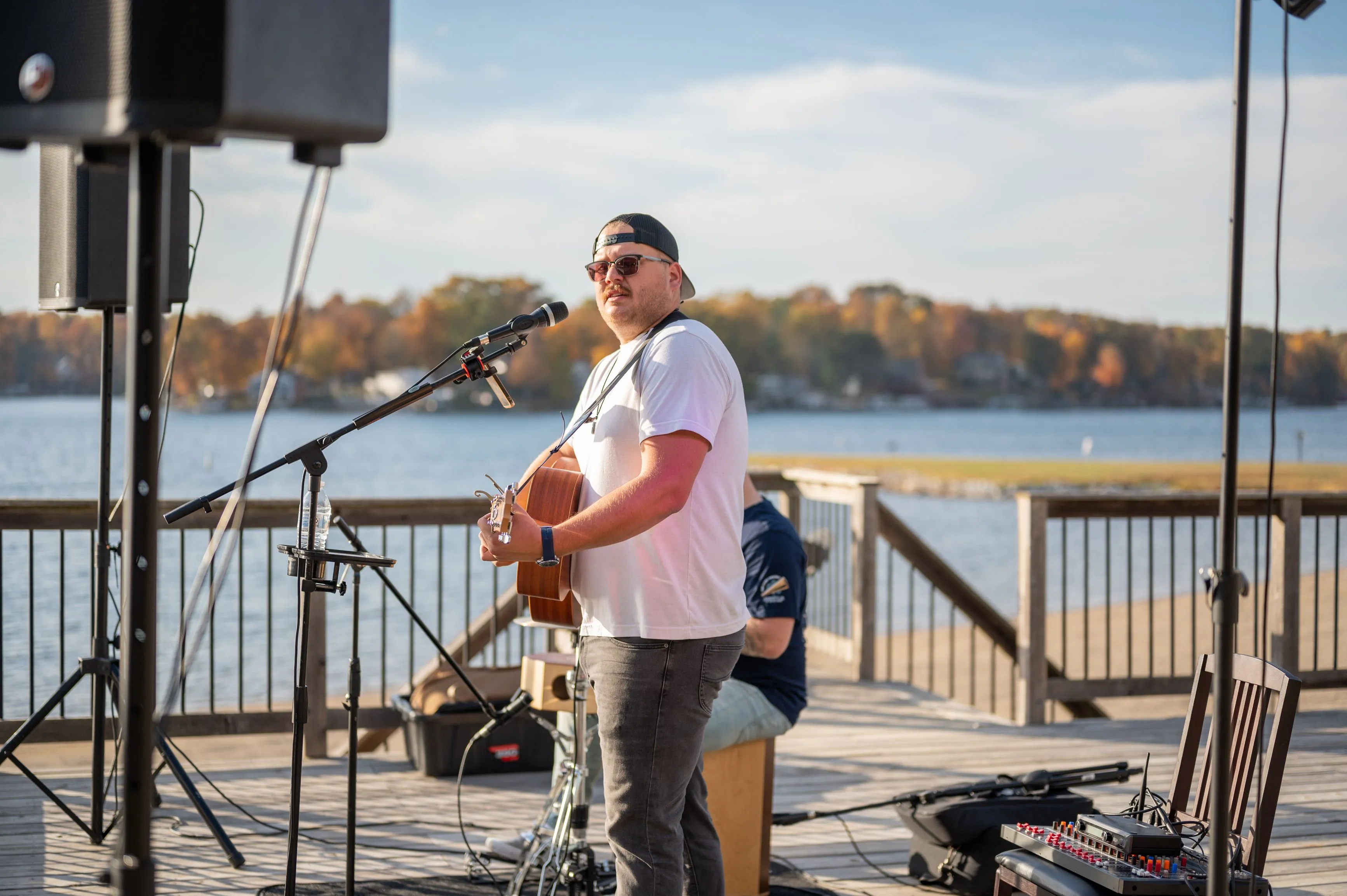Musician playing an acoustic guitar on a lakeside stage with microphone and speakers.