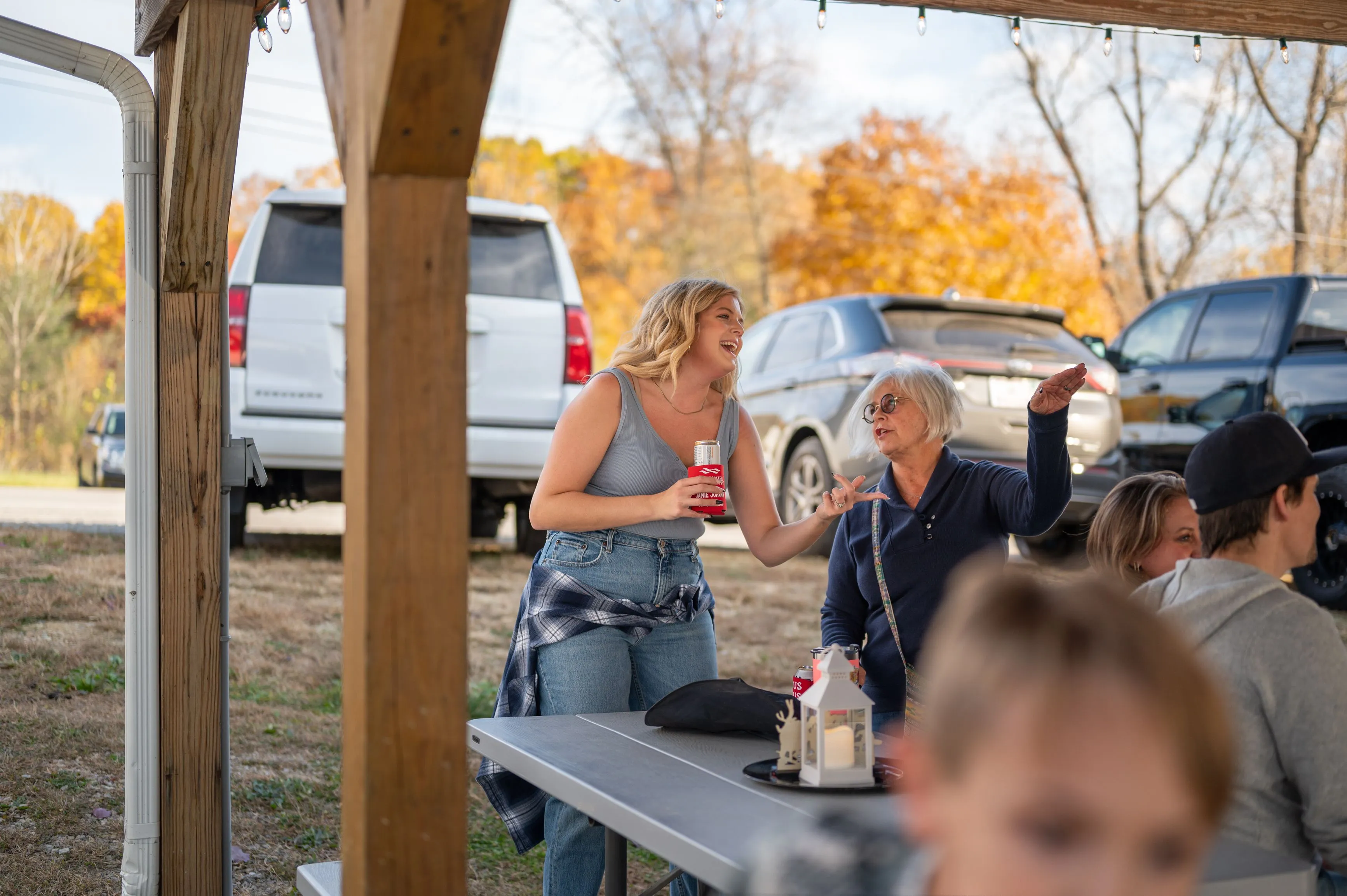 People socializing at an outdoor picnic area with vehicles parked in the background during autumn.