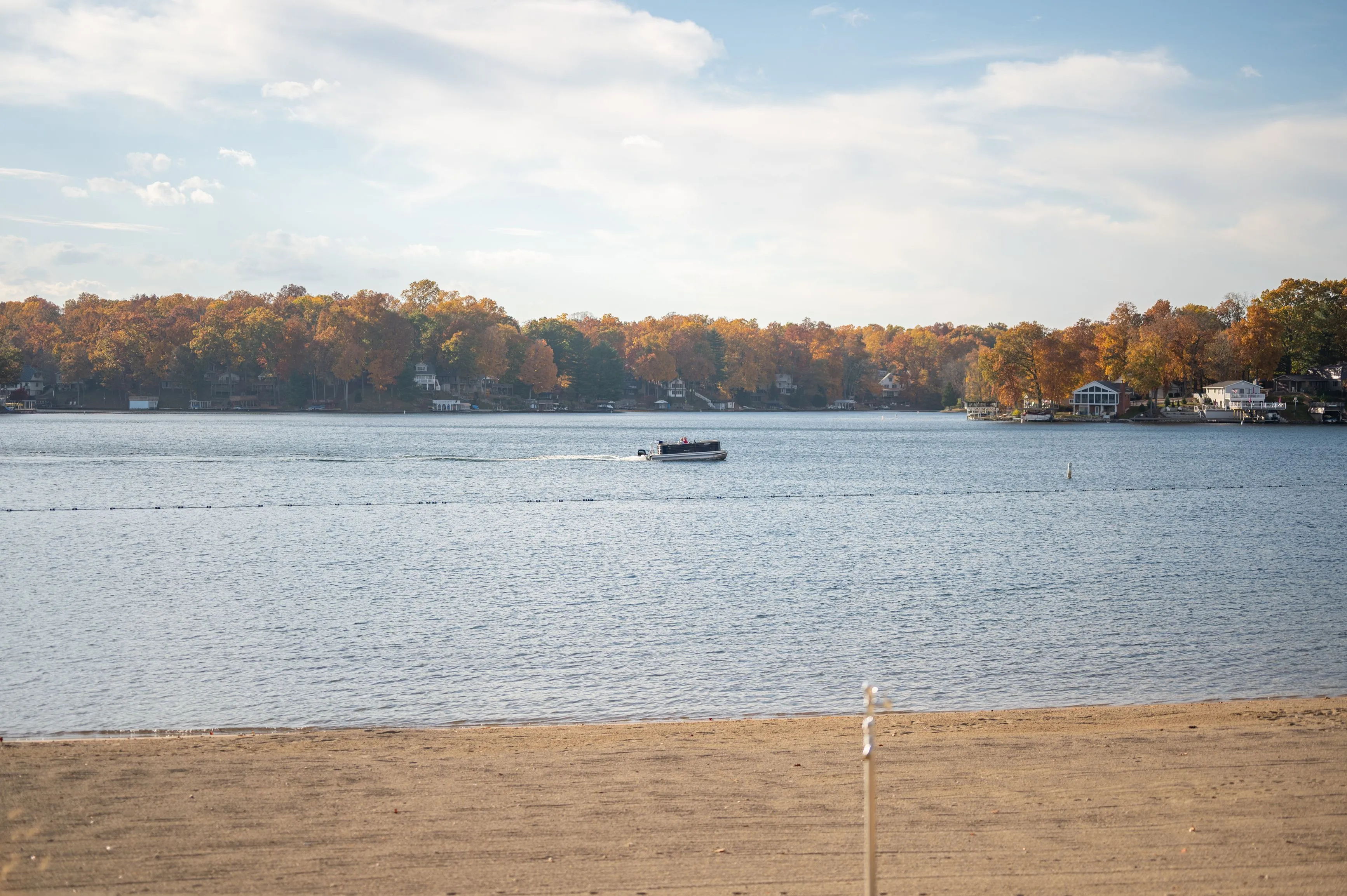 A tranquil lake with a boat in the distance and trees with autumn foliage on the far shore, viewed from a sandy beach.