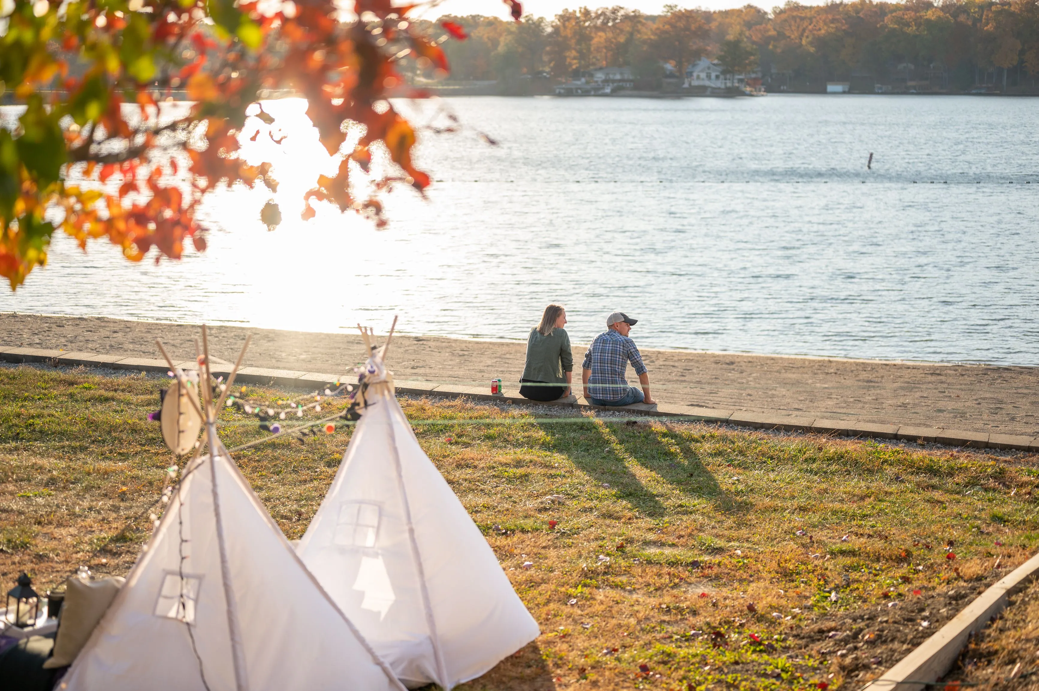Two people sitting by the lake with small white tents and autumn leaves in the foreground.