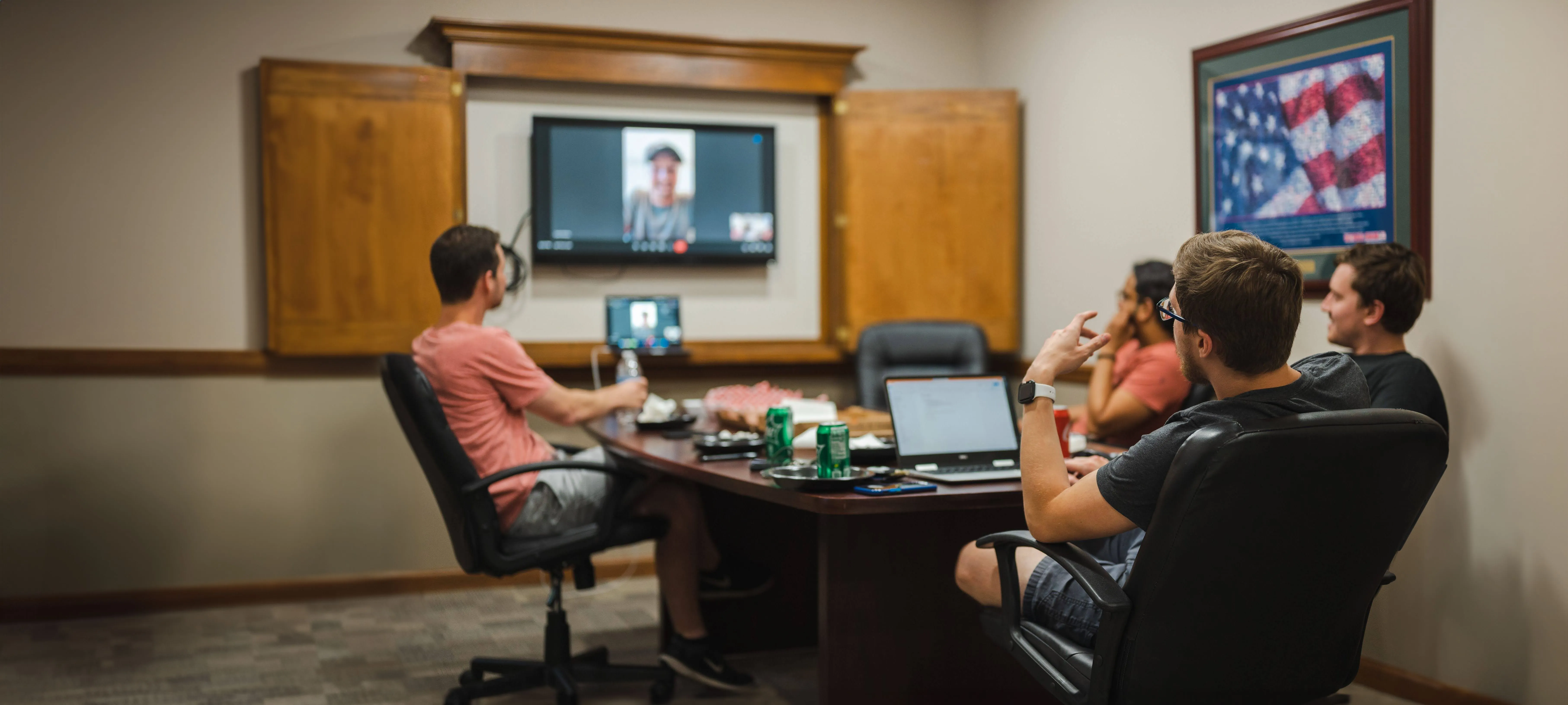 Group of people in a conference room engaging in a video call with a person displayed on a screen, with snacks and laptops on the table.