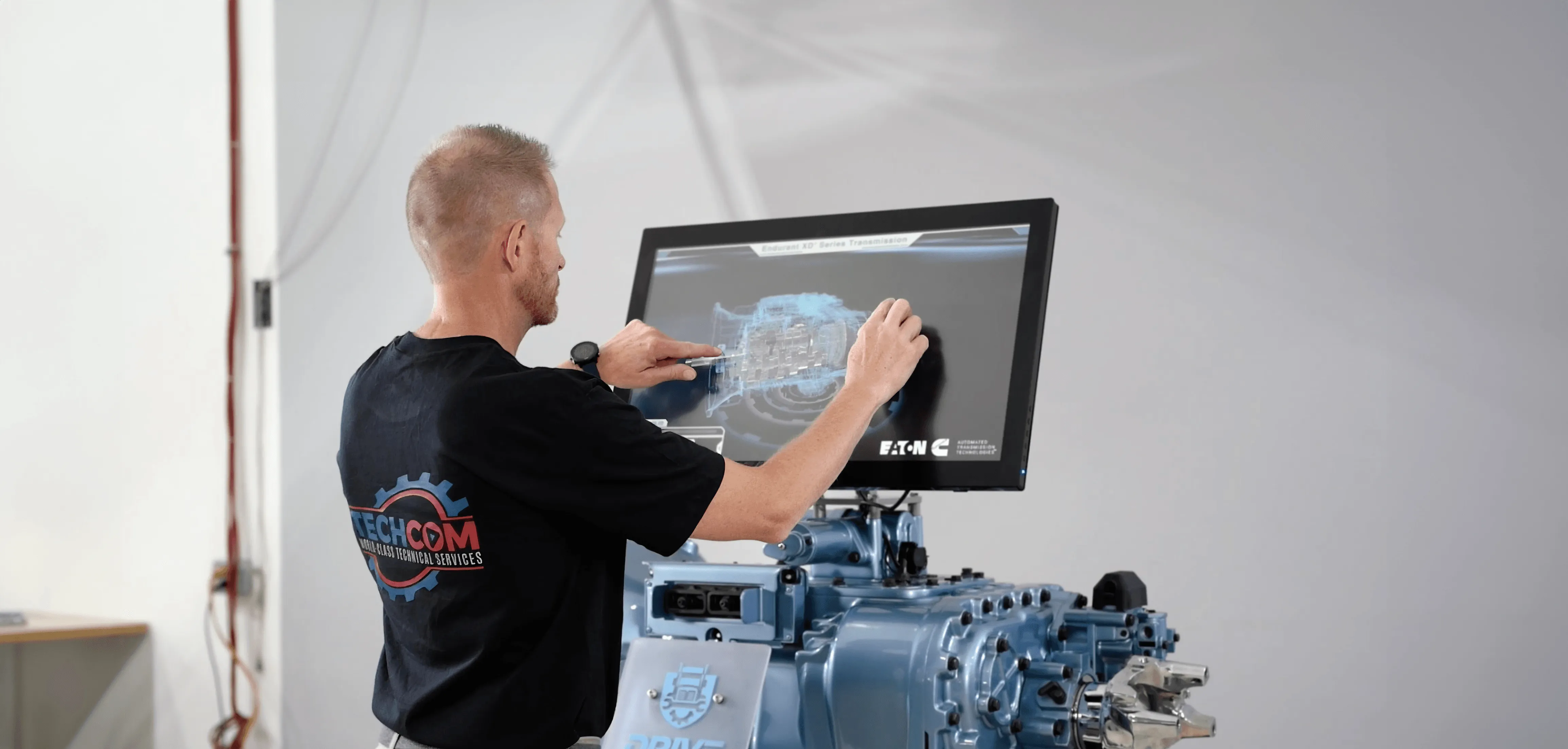 A technician interacting with a digital touchscreen displaying a 3D model of a transmission system with an actual engine component in the foreground.