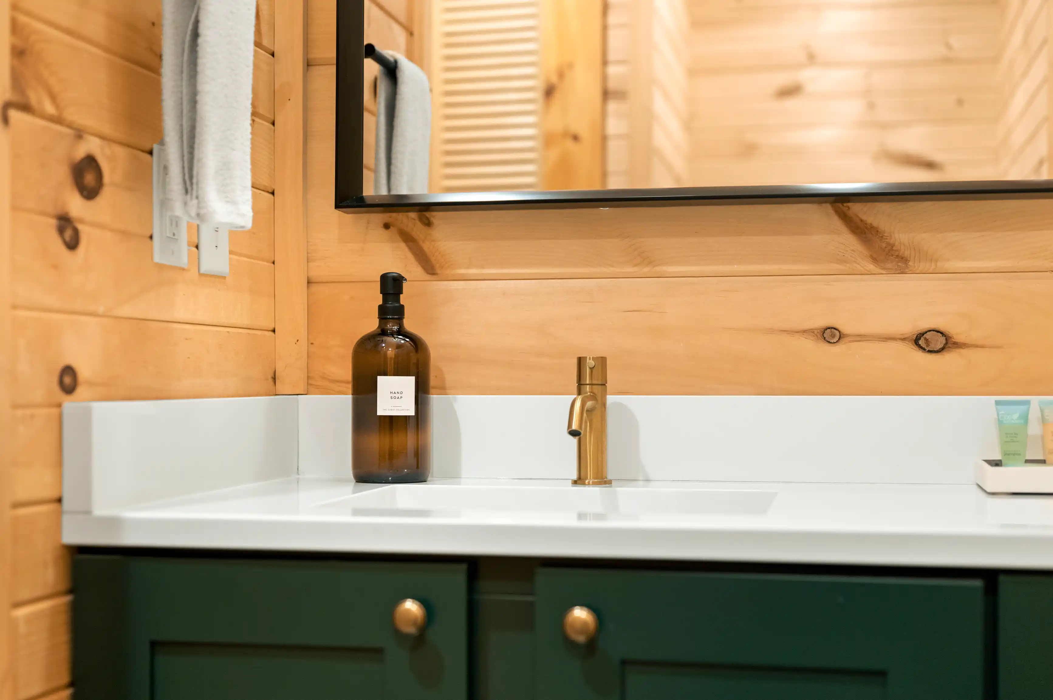 Modern bathroom interior with a white sink on a green vanity cabinet, amber glass soap dispenser, brass faucet, and mirror reflecting a wooden wall with a towel hanging.