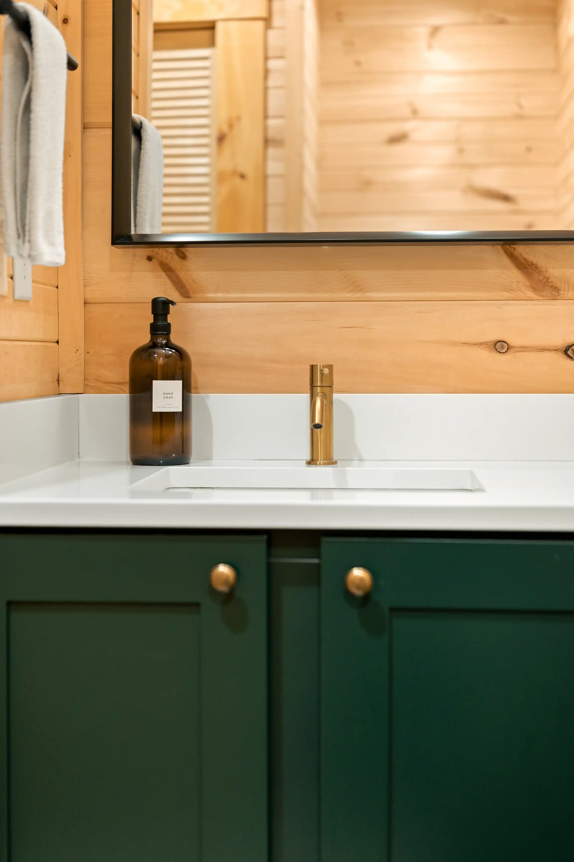 Modern bathroom sink with a white countertop, green cabinets, gold hardware, and a brown glass bottle labeled "HAND SOAP"; reflection of wooden interior visible in the mirror above.