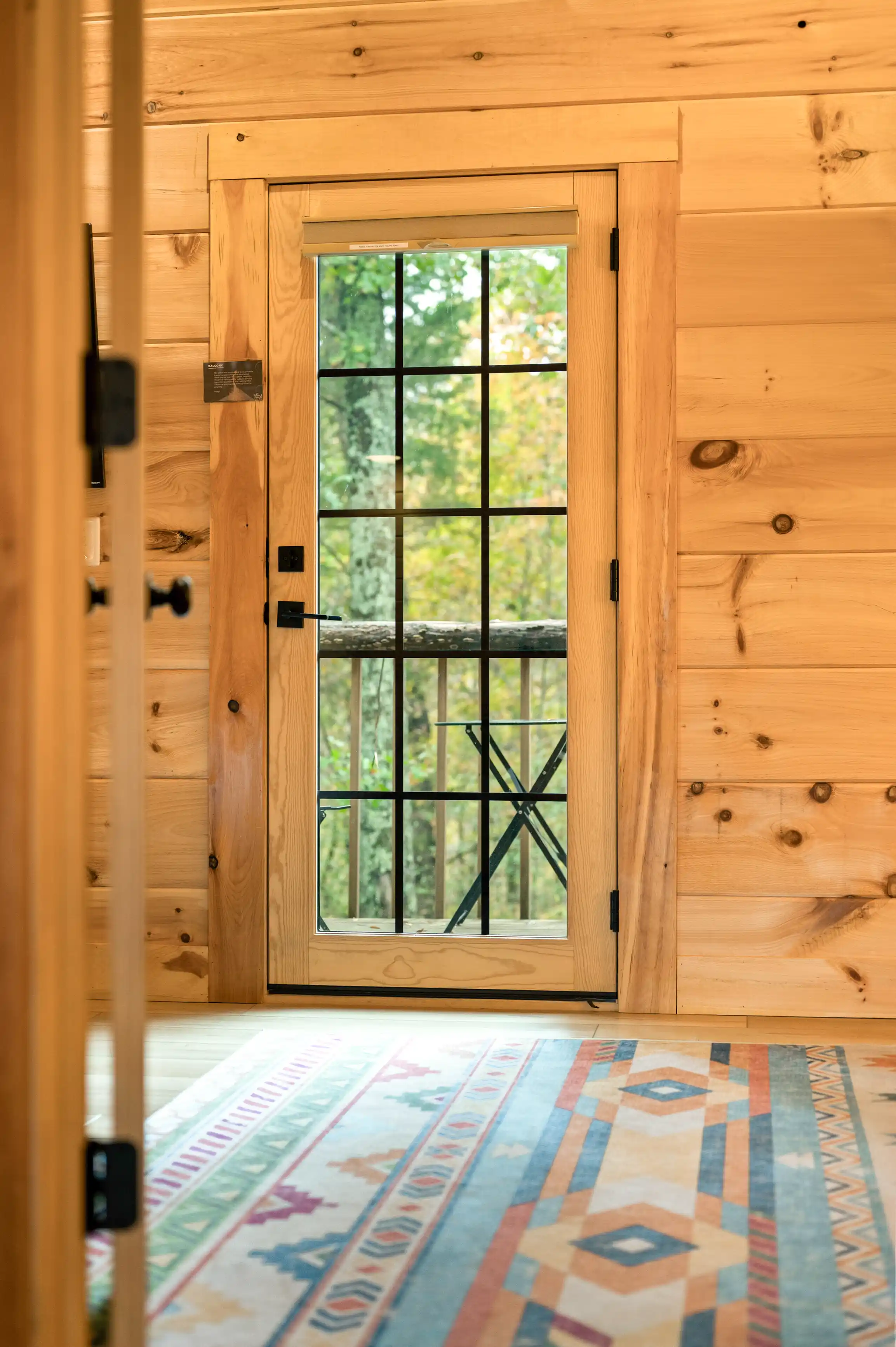 Wooden door with glass panes set in a natural wood-finished room, leading to an outdoor area, with a colorful patterned rug in the foreground.