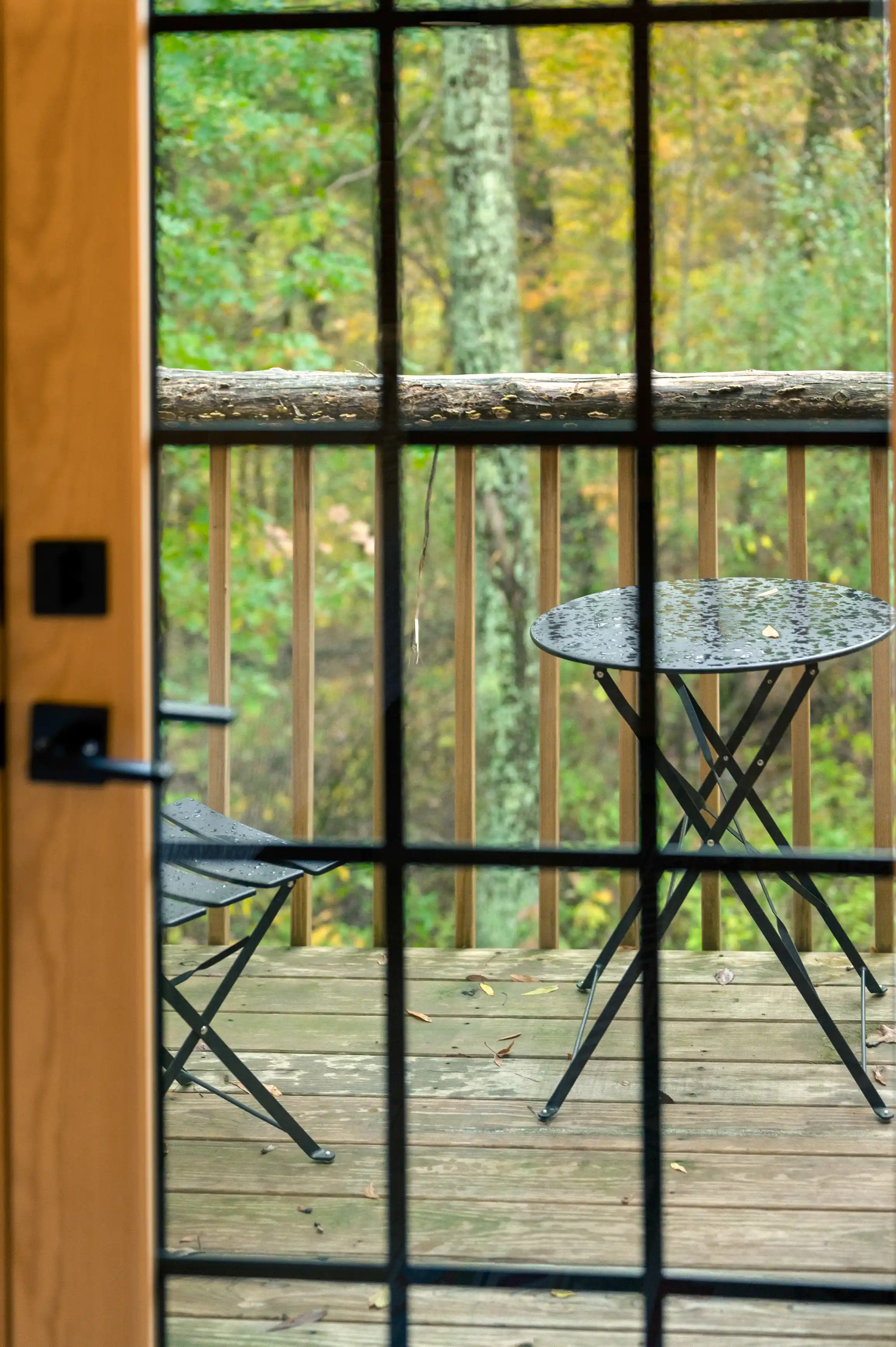 View from a window showing a wet bistro table and chair on a wooden deck overlooking a forest.