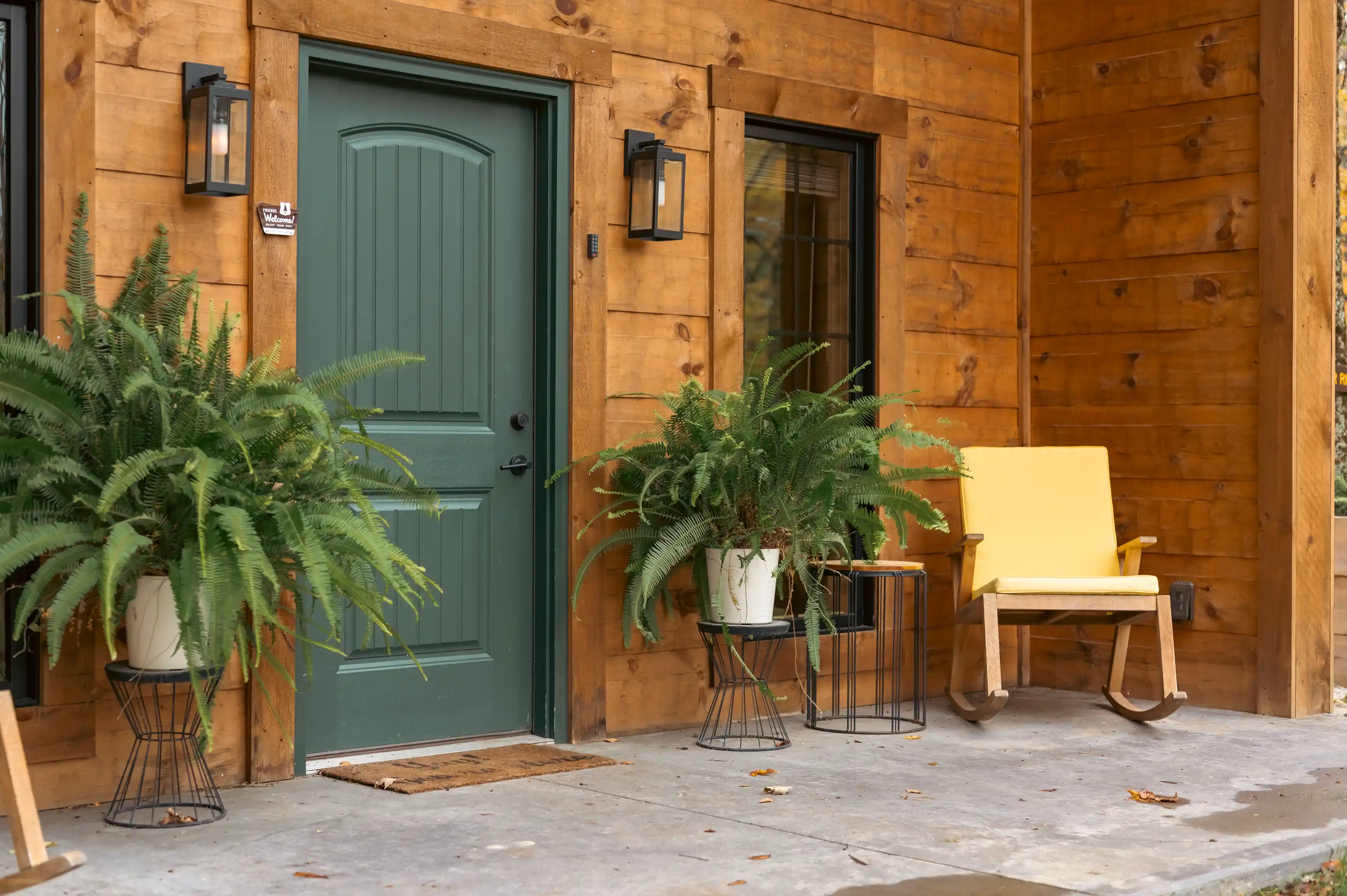 Cozy house entrance with a green door flanked by ferns and an inviting yellow chair on the porch.