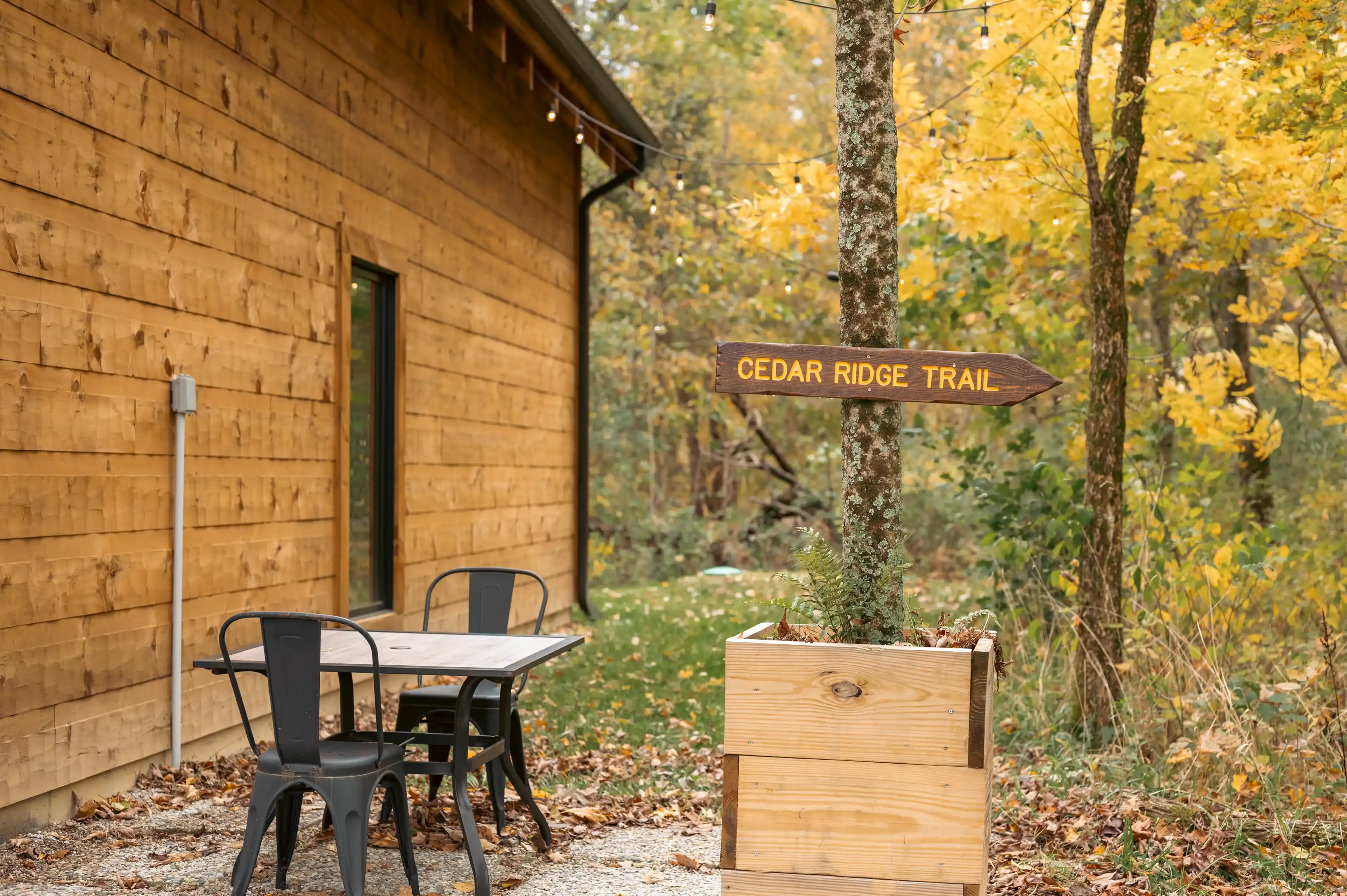 Outdoor setting with a "CEDAR RIDGE TRAIL" signpost near a cabin, with a table and chairs on gravel, surrounded by autumn trees.