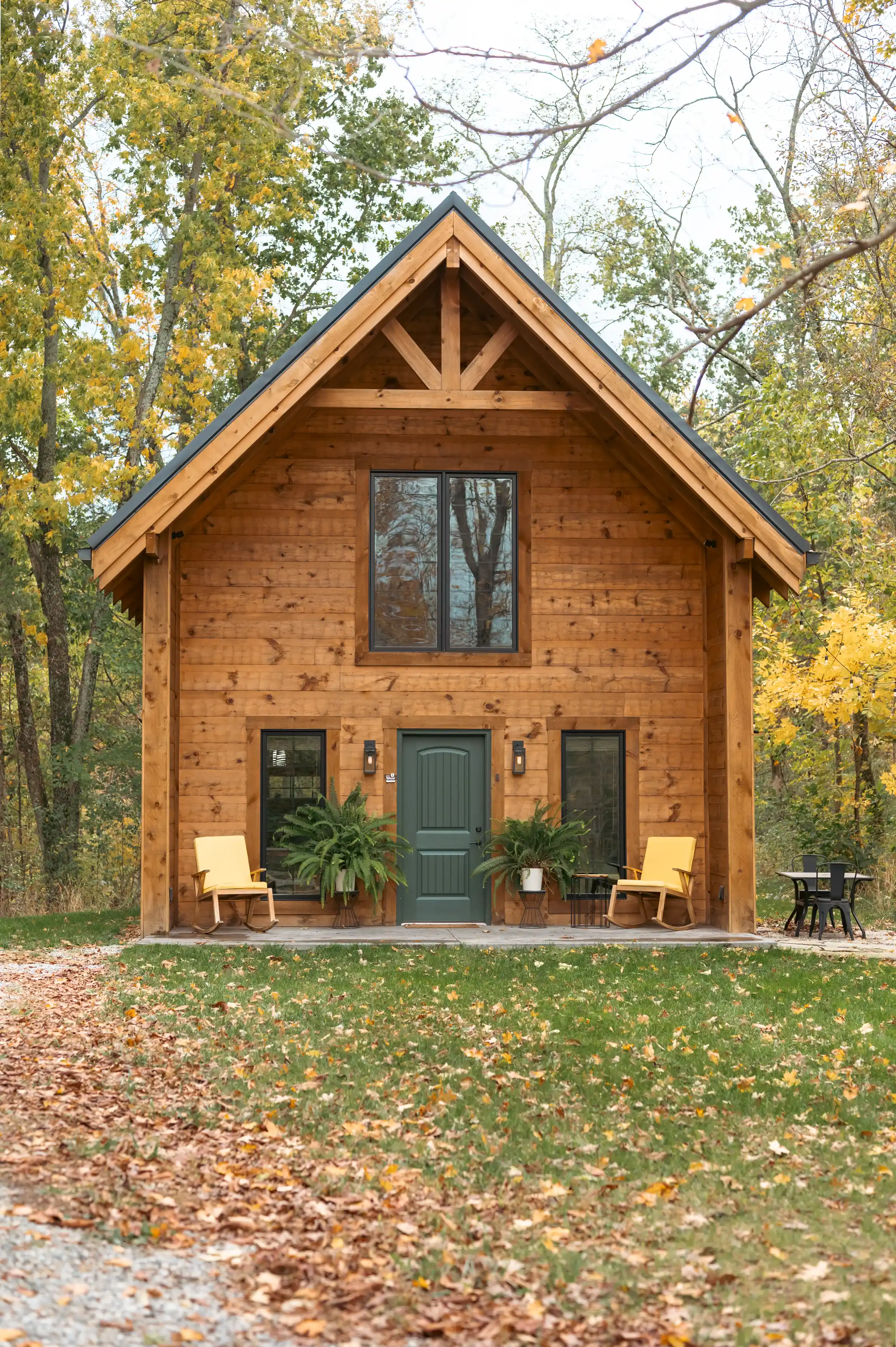A wooden cabin with a green door, large front window, and a porch with chairs surrounded by trees with autumn leaves.