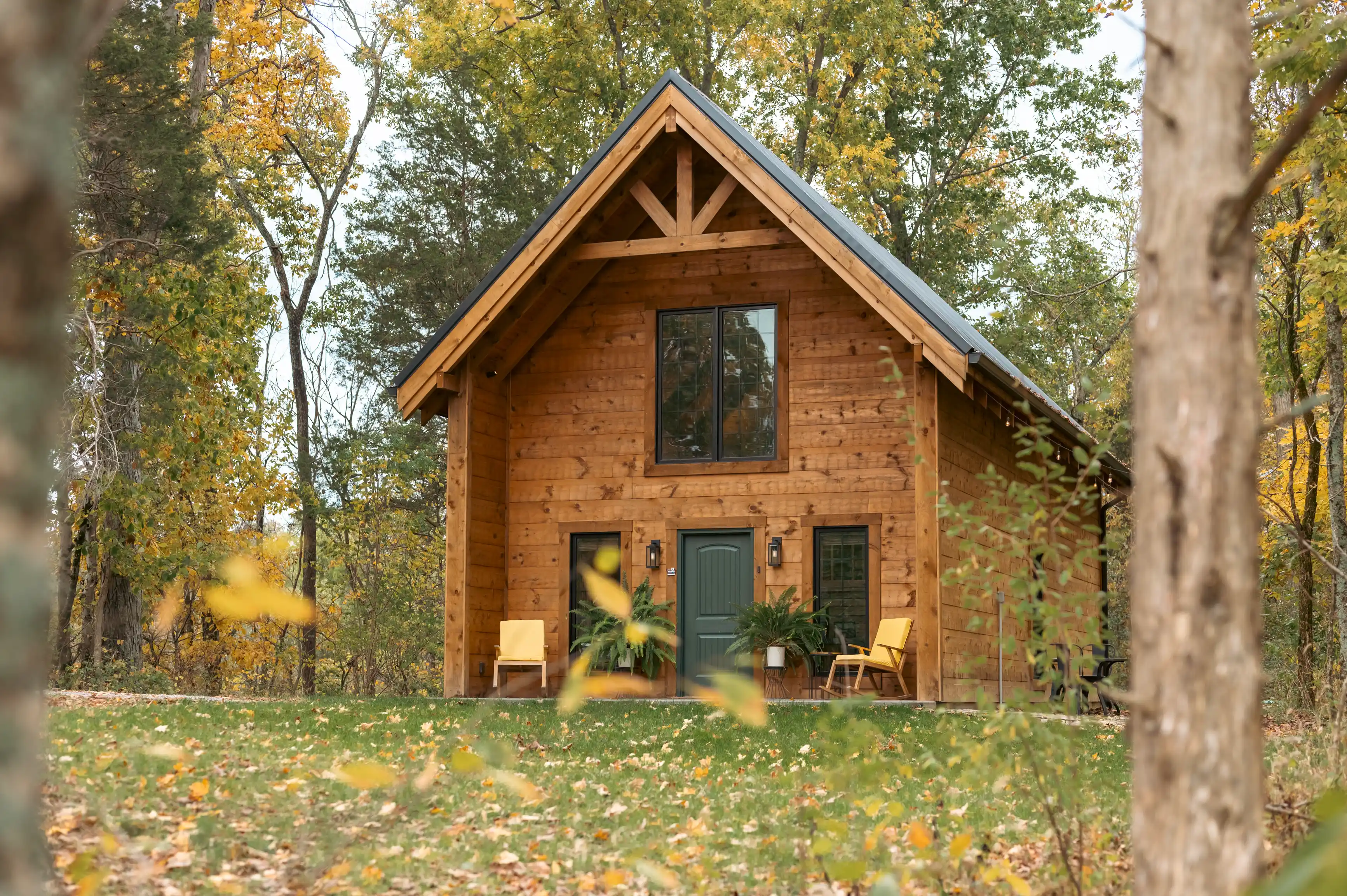 A wooden cabin with a green door nestled among autumn trees, featuring a gabled roof and large front window, with yellow chairs on the porch.