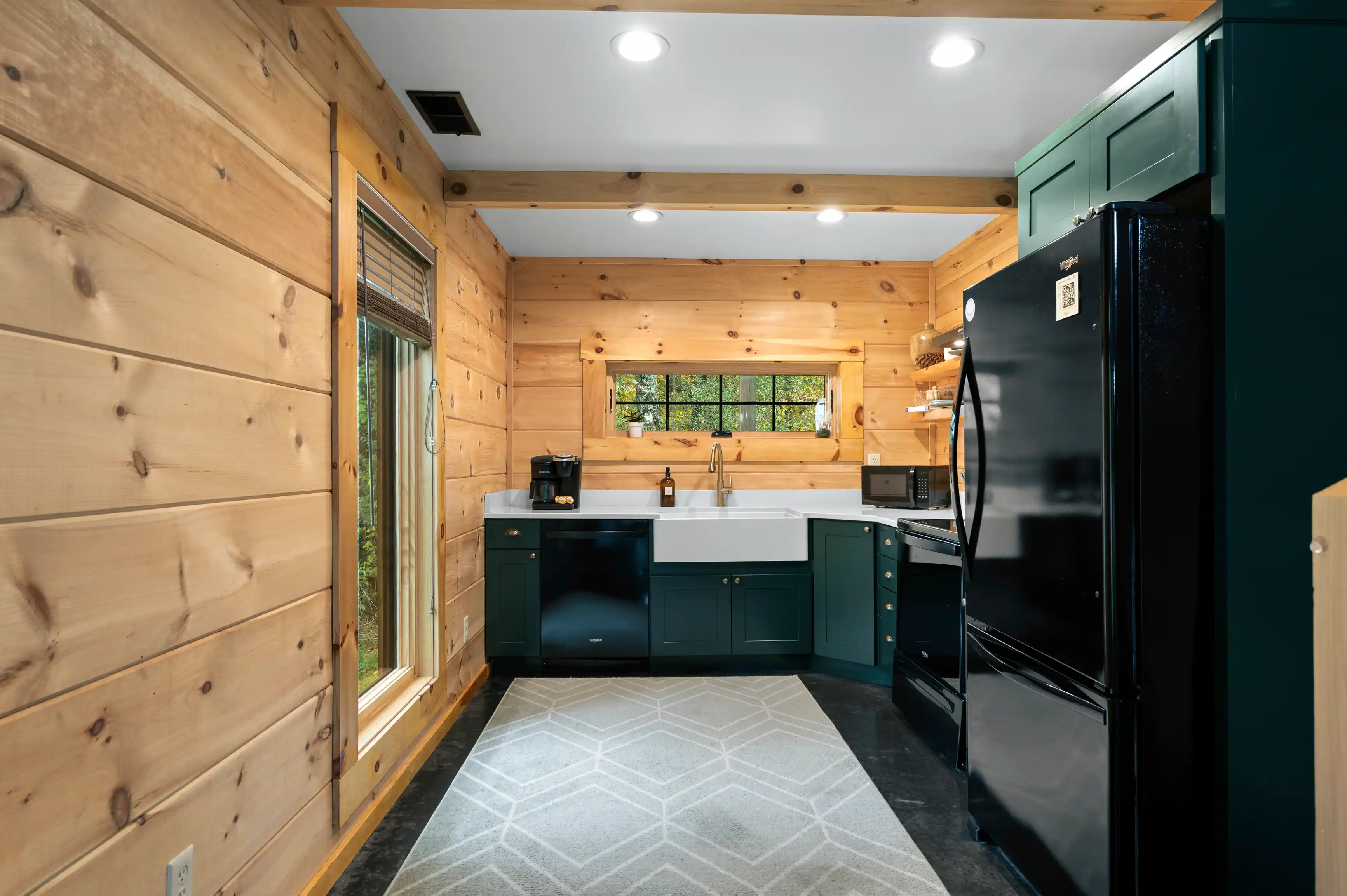Interior view of a kitchen with dark green cabinets, wooden walls, and modern appliances, featuring a geometric patterned rug on the floor.