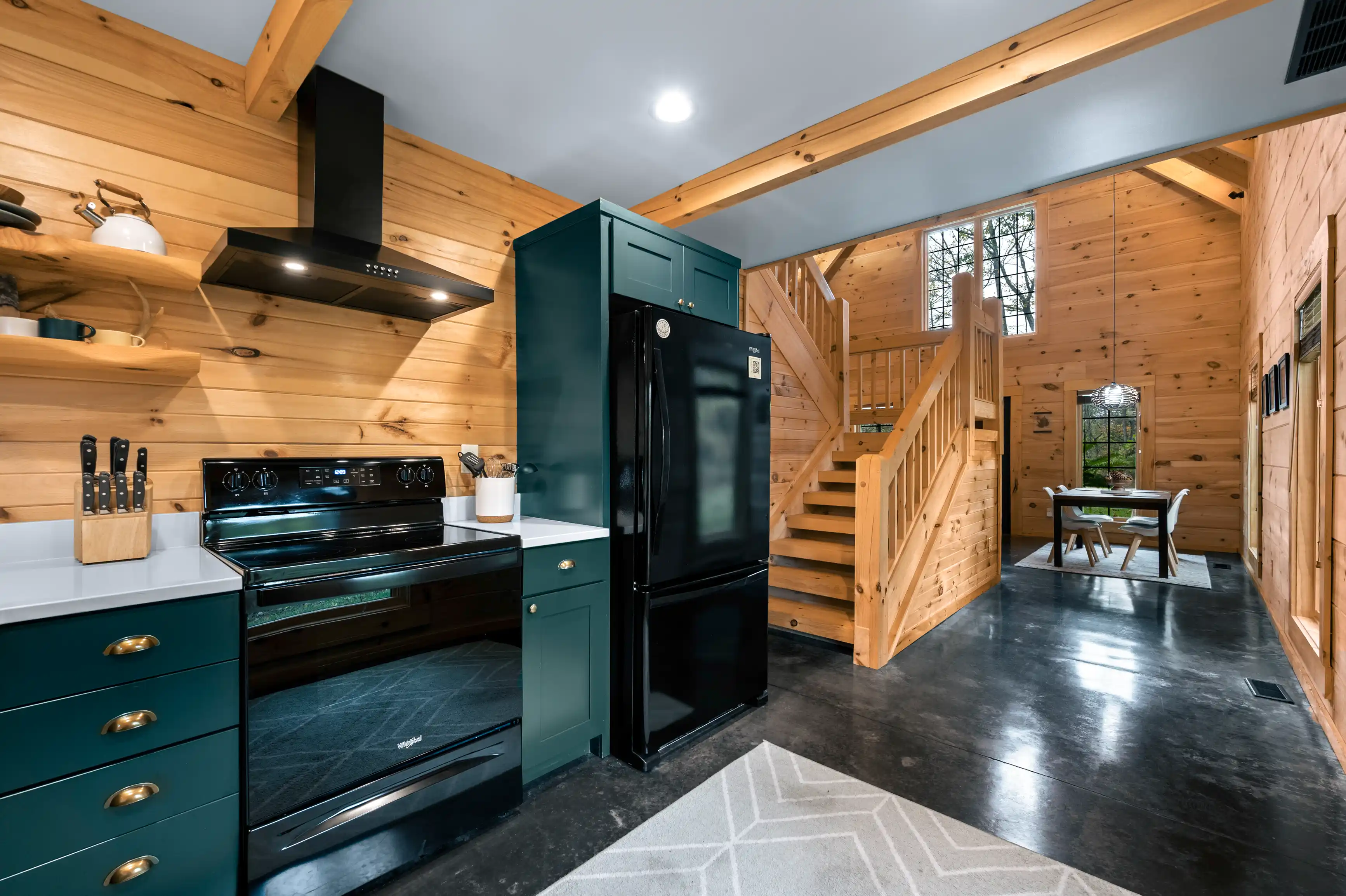 Modern kitchen interior with dark green cabinets and black appliances contrasted against natural wood walls and ceiling, featuring a staircase leading to an upper level.