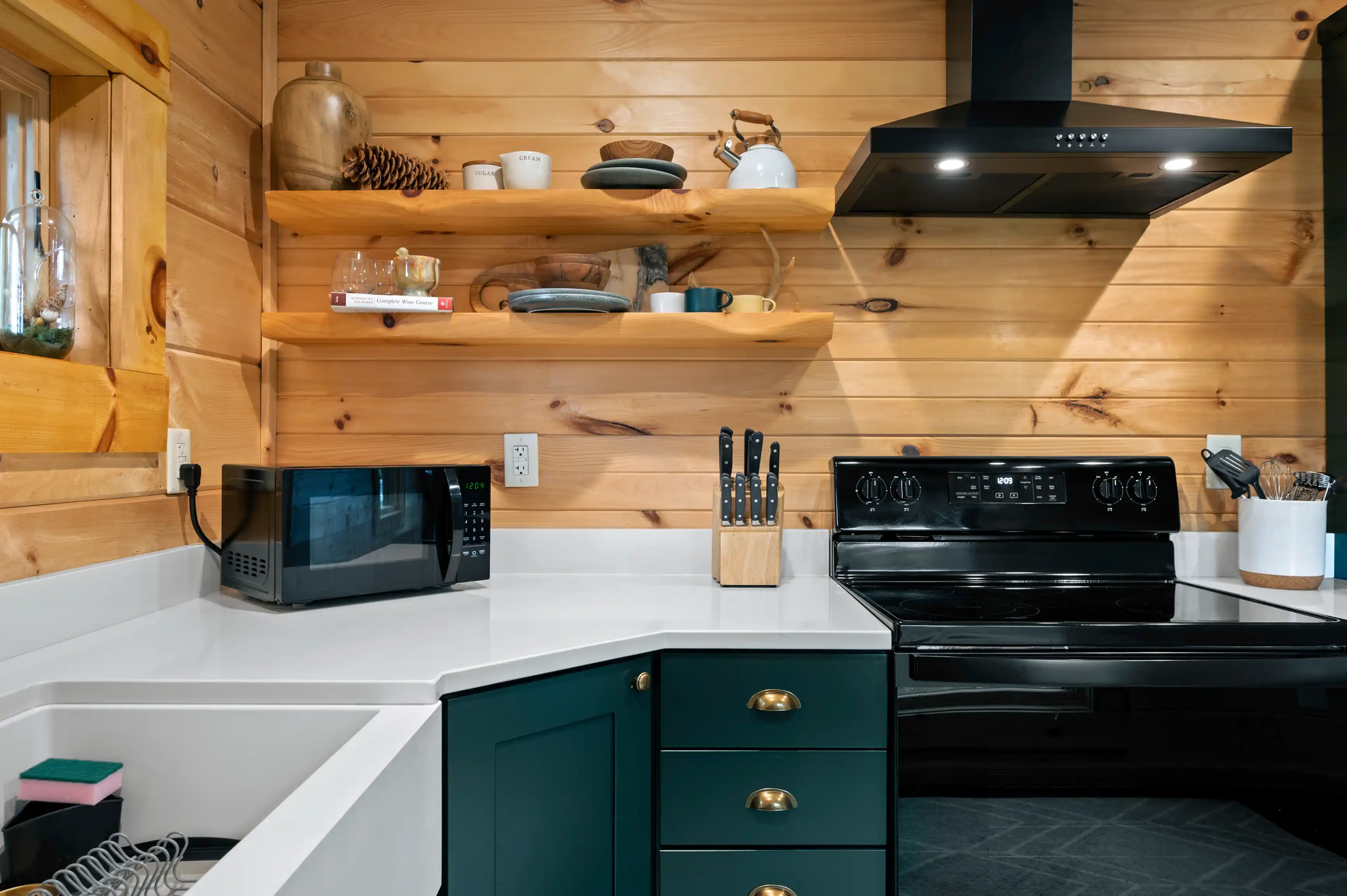 Modern kitchen interior with wooden walls and green cabinets, featuring a microwave, a stove, floating shelves with dishes, and cooking utensils.