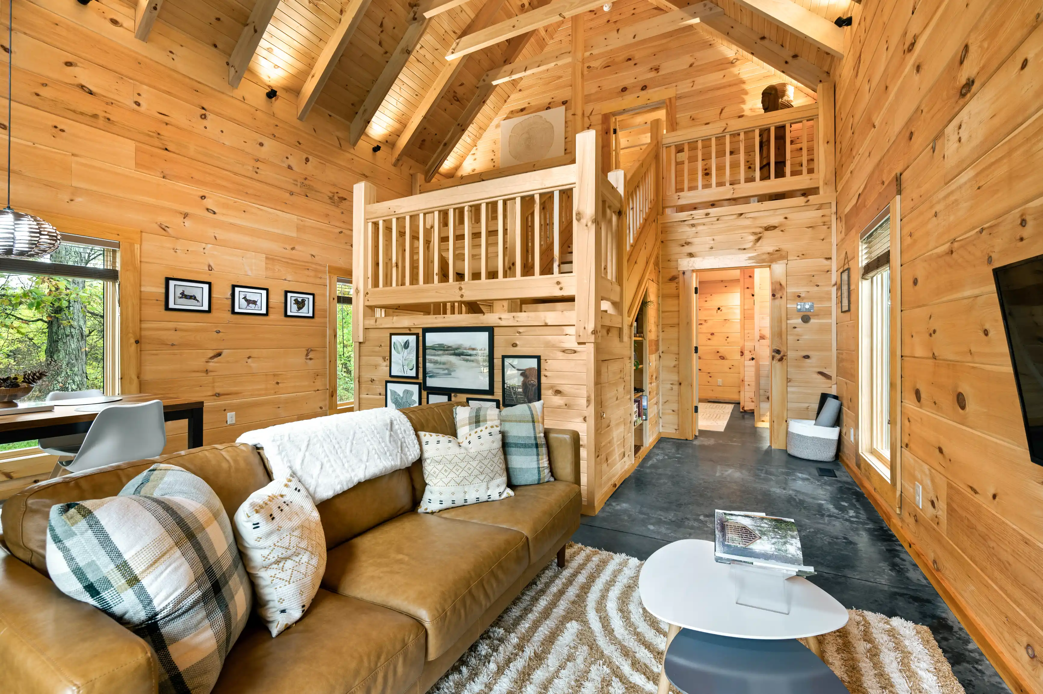 Interior of a cozy cabin living room with wooden walls, a high ceiling, loft area, large windows, plush sofa with pillows, and modern decorations.