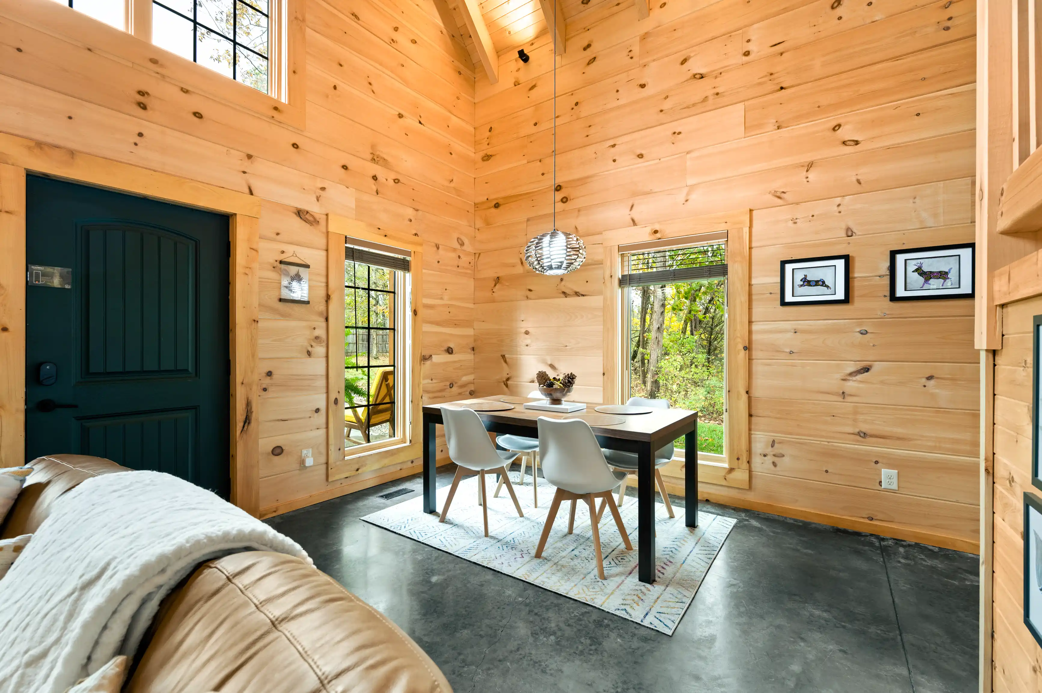 Cozy wooden cabin interior with a dining area, modern furniture, and large windows showing lush greenery outside.