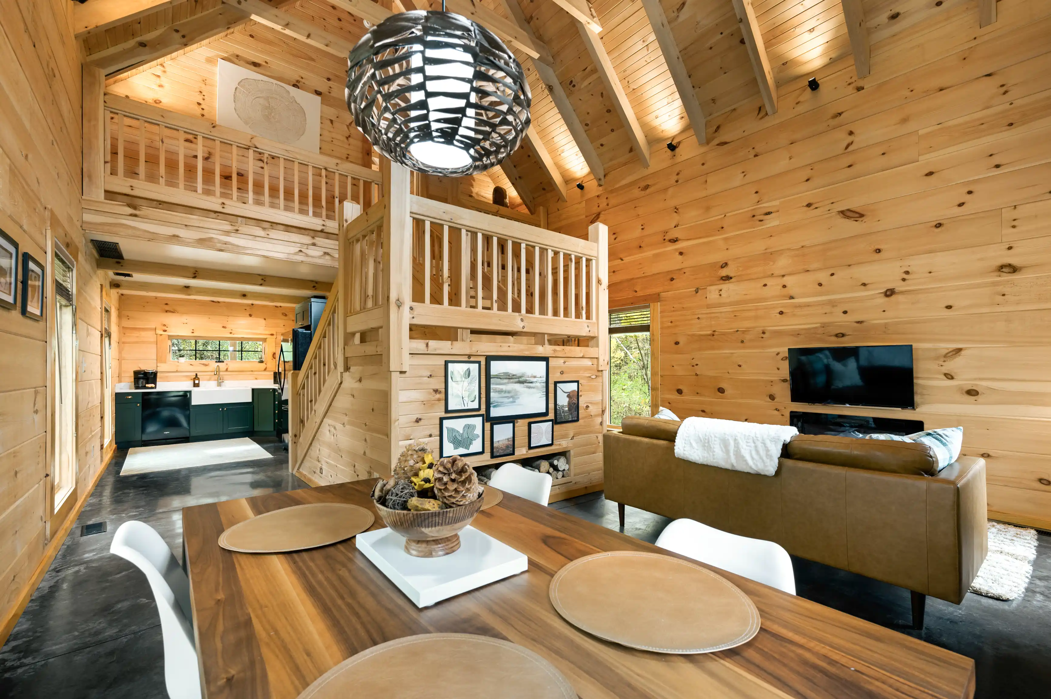 Interior of a cozy wooden cabin with a modern kitchen, a living room with a flat-screen TV, comfortable furniture, a loft area accessed by stairs, and a distinctive hanging light fixture.
