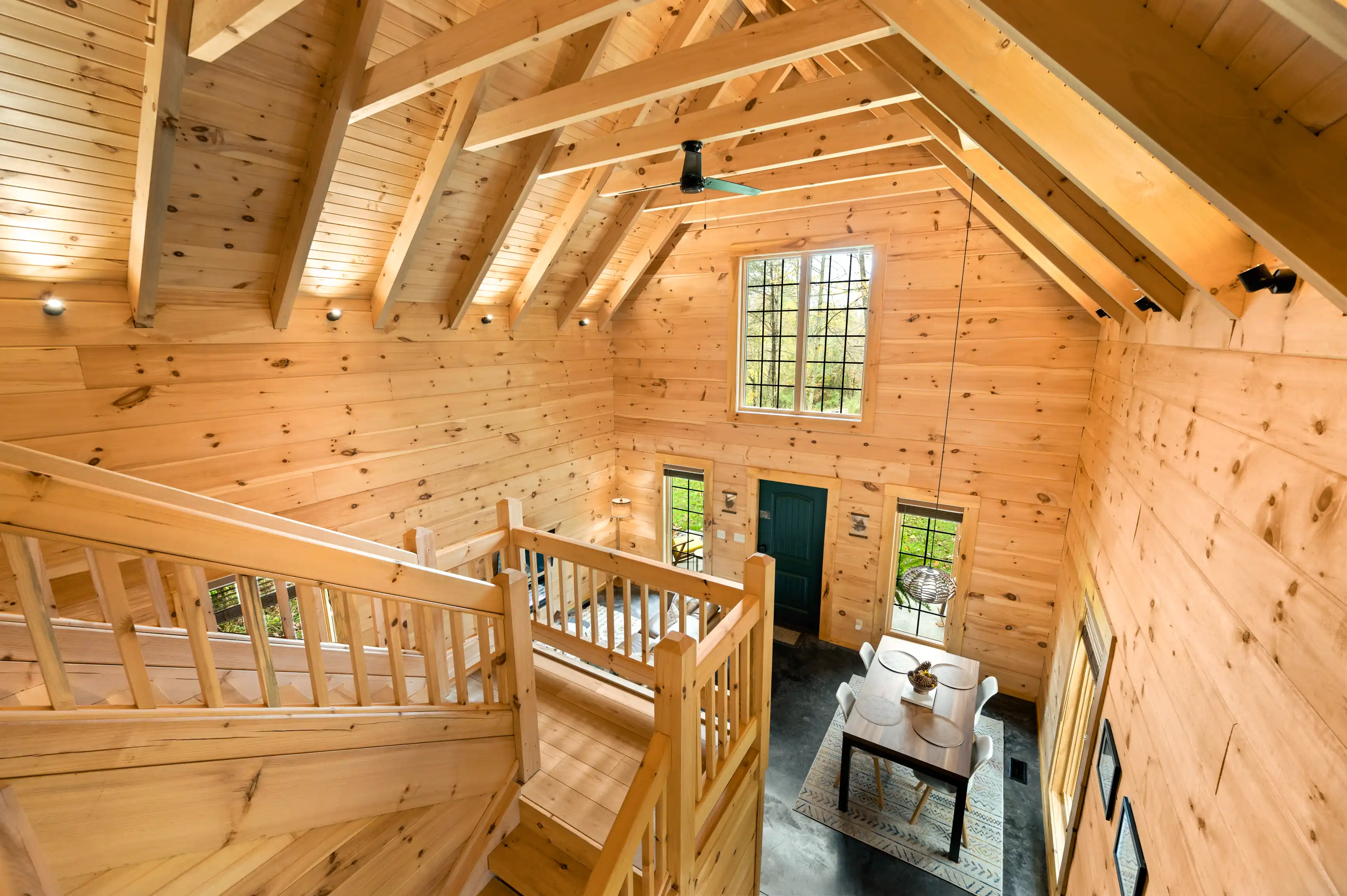 Interior view of a spacious cabin with wooden walls and rafters, featuring large windows and a rustic staircase leading to an upper floor.